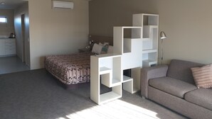 A bedroom with a bed, a sofa, a desk with shelves, a lamp, and a kitchenette in the background. - Tinwald Motels (Ashburton)