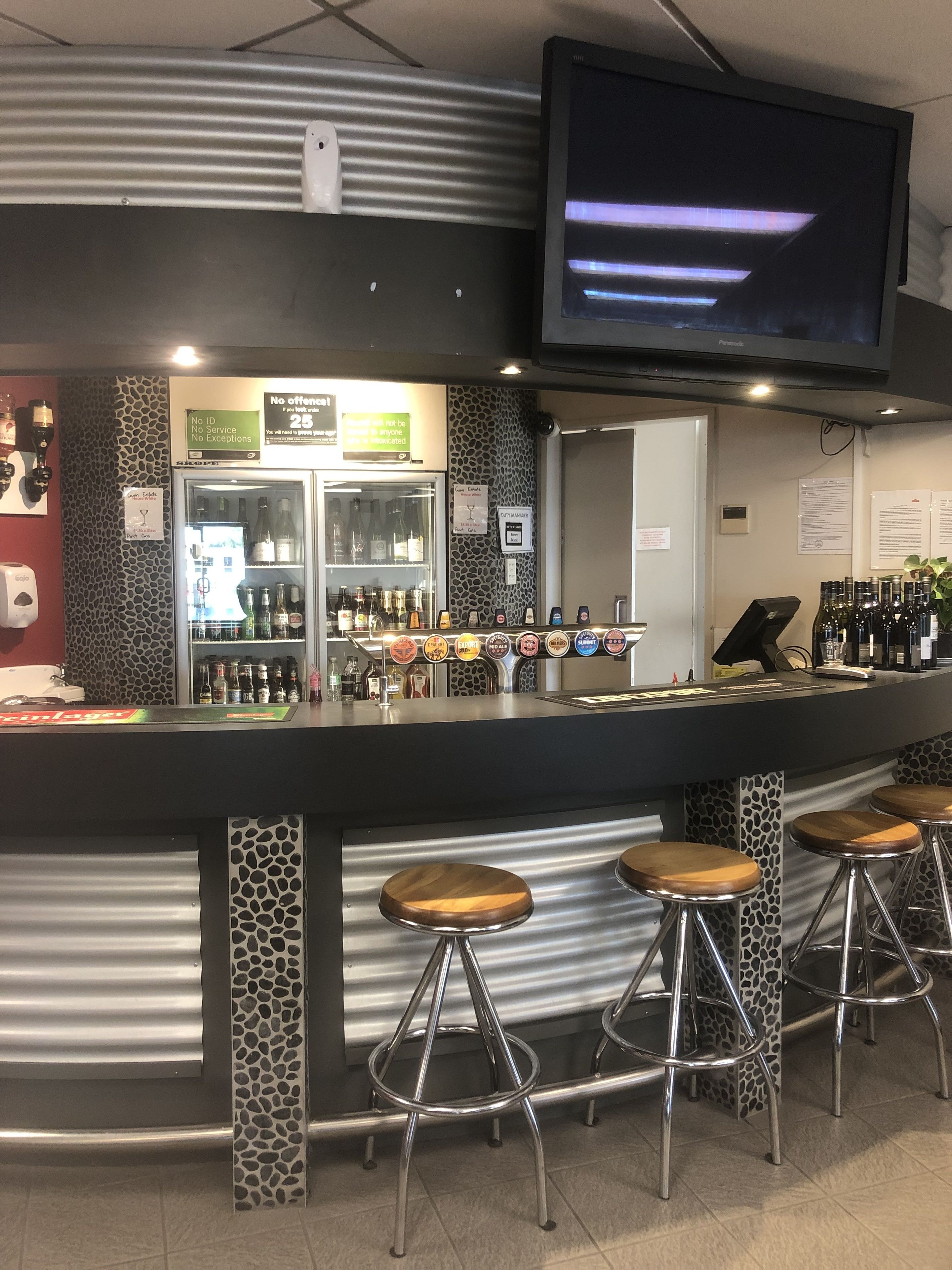 Un área de bar con una barra, taburetes, un refrigerador lleno de bebidas y un televisor montado en la pared.