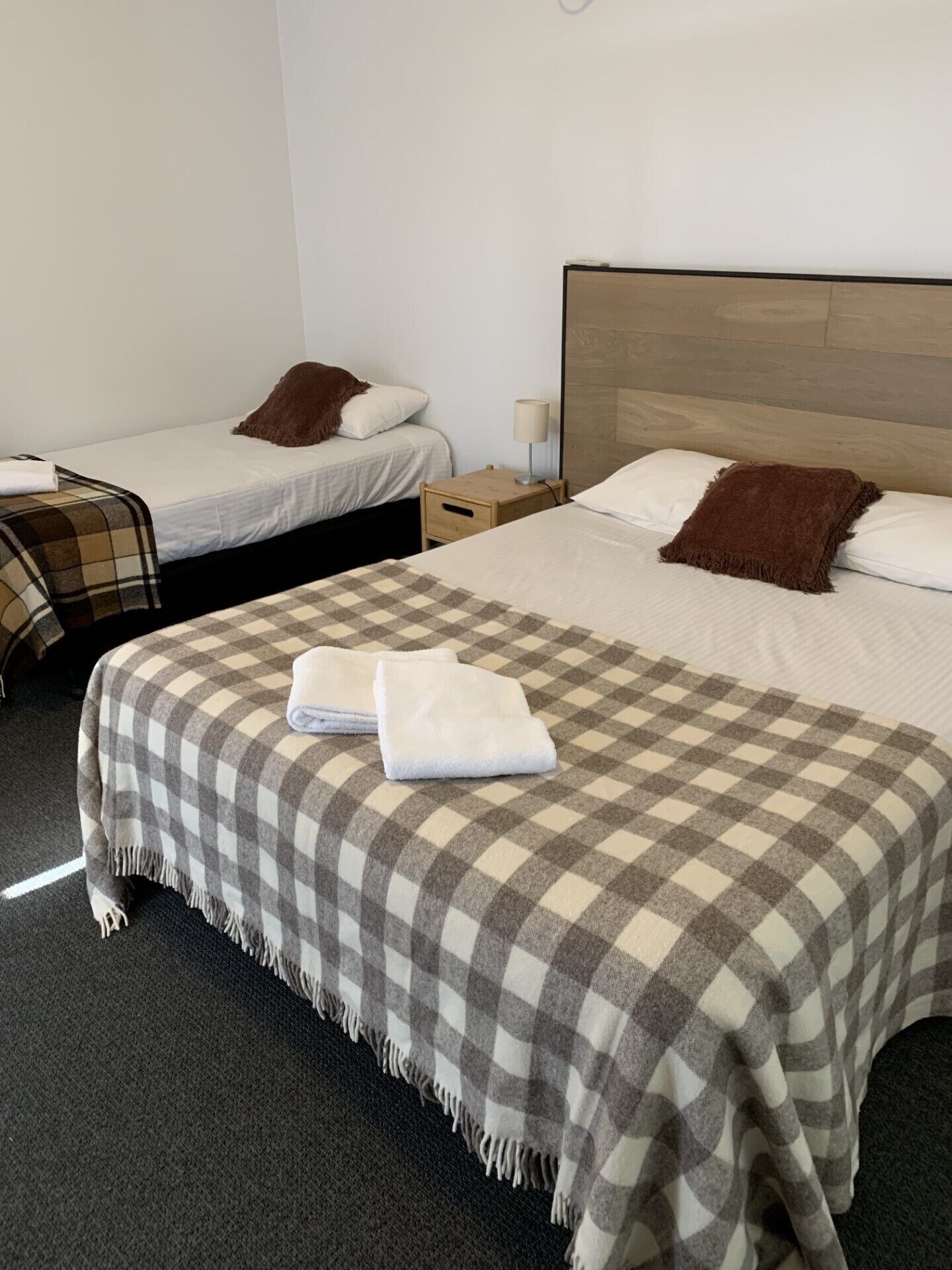Una habitación de hotel con dos camas, cada una con almohada y manta. Entre las camas, una mesita de noche con lámpara.