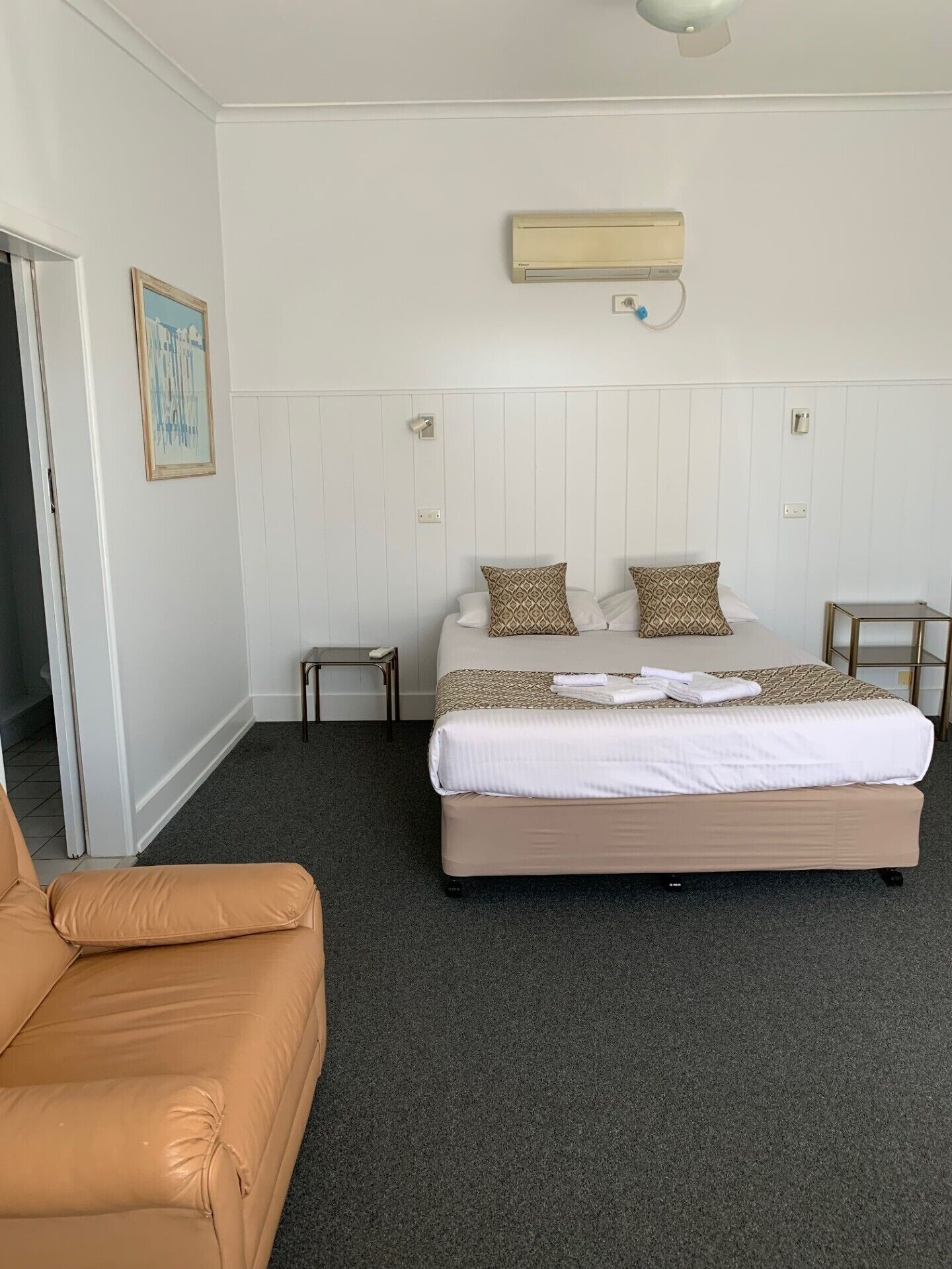 A hotel room with a large bed, a small table, a chair, and a wall-mounted air conditioning unit.