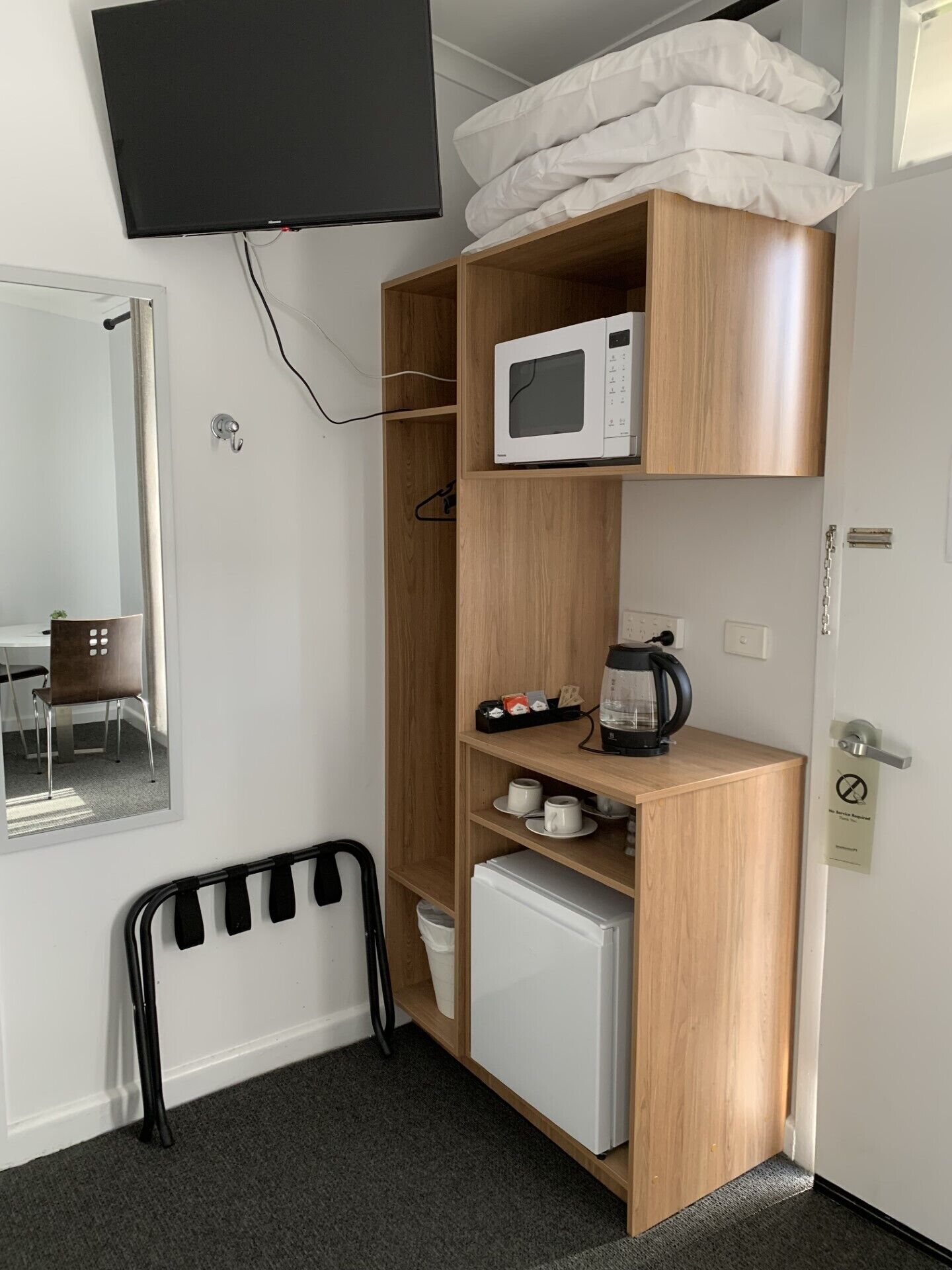 Ein kleiner Raum mit einem Holzschrank, der eine Mikrowelle, einen Wasserkocher und eine kleine Kühlbox beherbergt. Ein Sessel und ein Bett mit Kissen sind ebenfalls vorhanden.