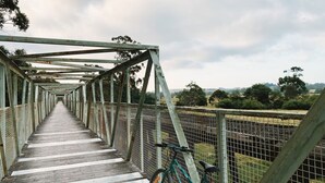 A long pedestrian bridge with a metal framework and wooden planks, leading to a fenced area with trees in the background. - Meeniyan Motel (Meeniyan)