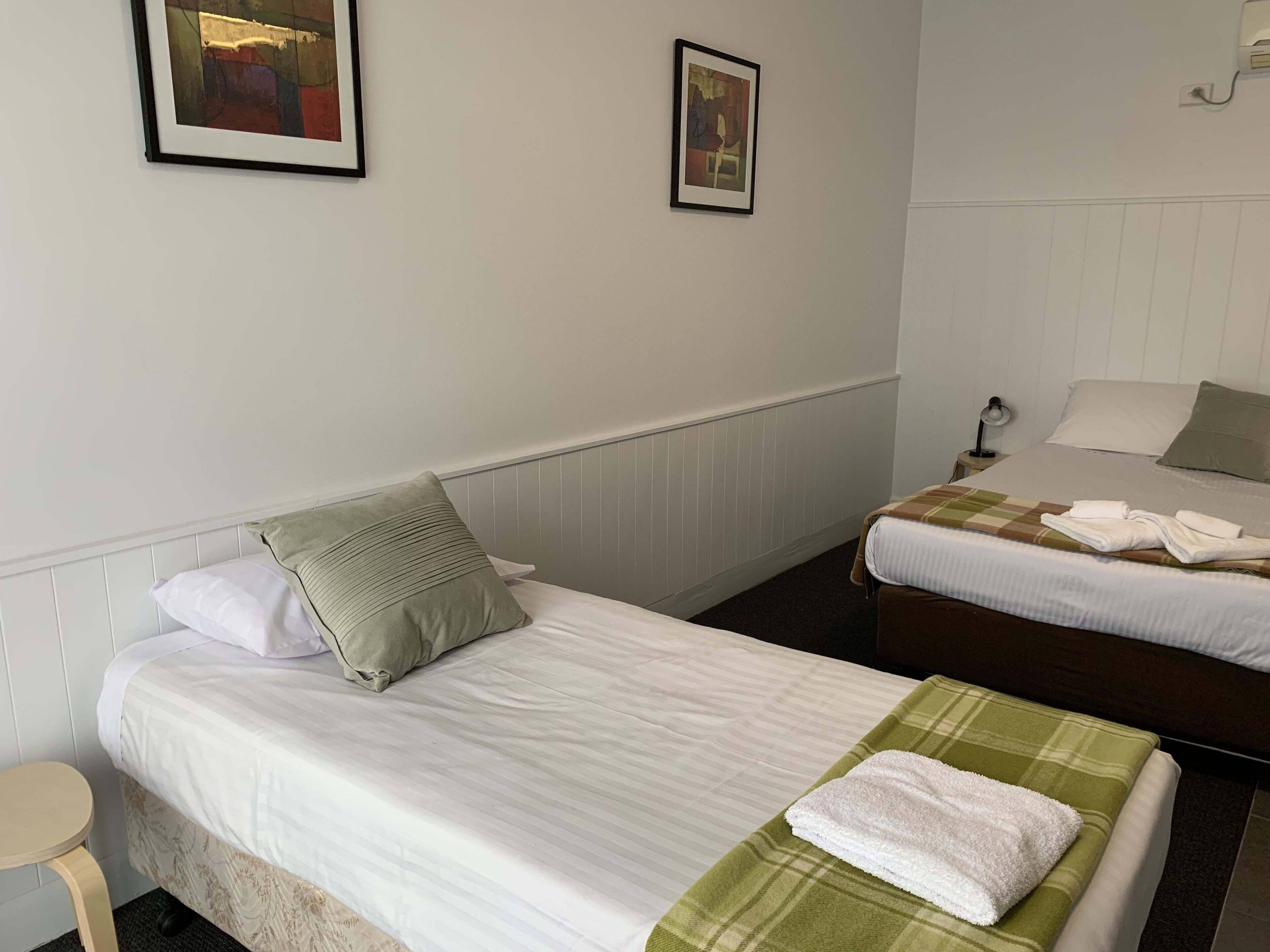 A hotel room with two single beds, a small table, and a wall-mounted lamp.