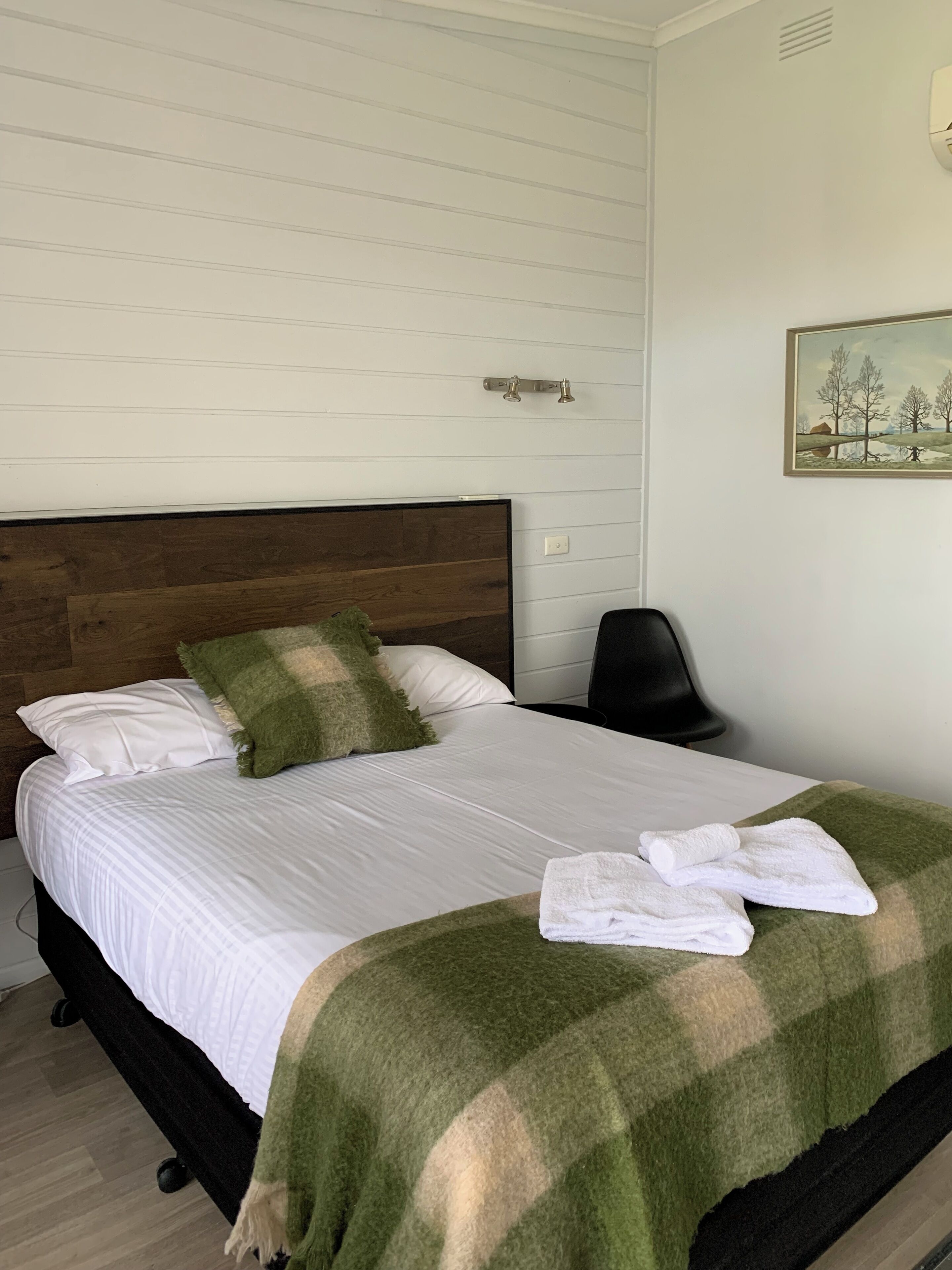A neatly made bed with white linens, a green and beige checkered blanket, and two pillows. There is a painting on the wall and a chair to the right of the bed.