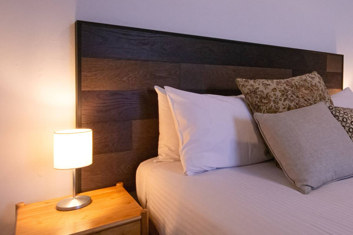 Una cama bien hecha con almohadas blancas y estampadas, una mesita de noche de madera con una lámpara y un cabecero con diseño de paneles de madera oscura.