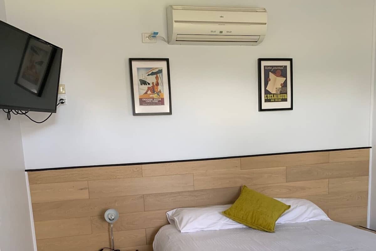 Habitación de hotel con una cama, una mesita de noche con lámpara, aire acondicionado y dos cuadros en la pared.