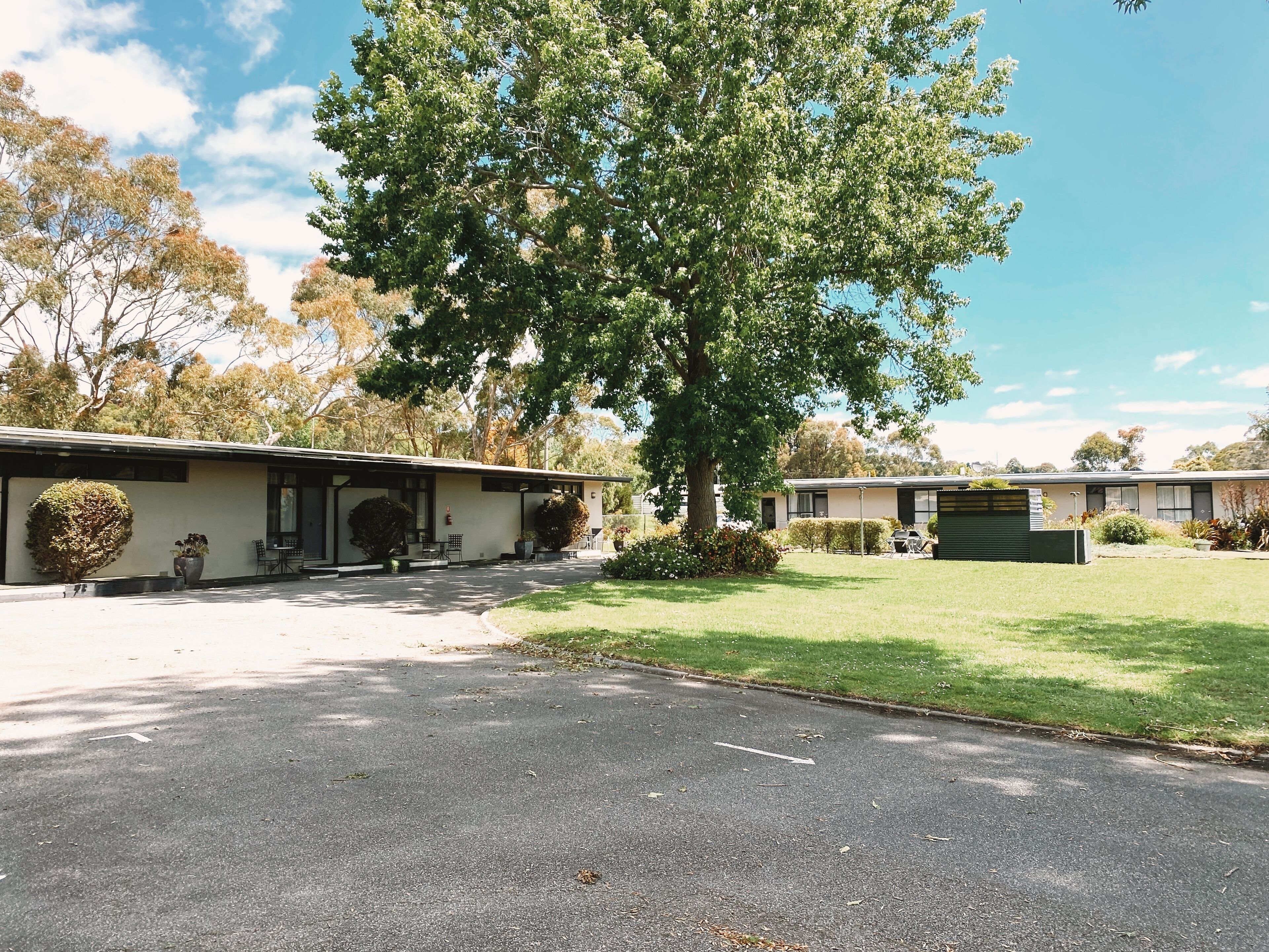 A motel with a large tree in the front yard, a covered outdoor seating area, and a well-maintained lawn.