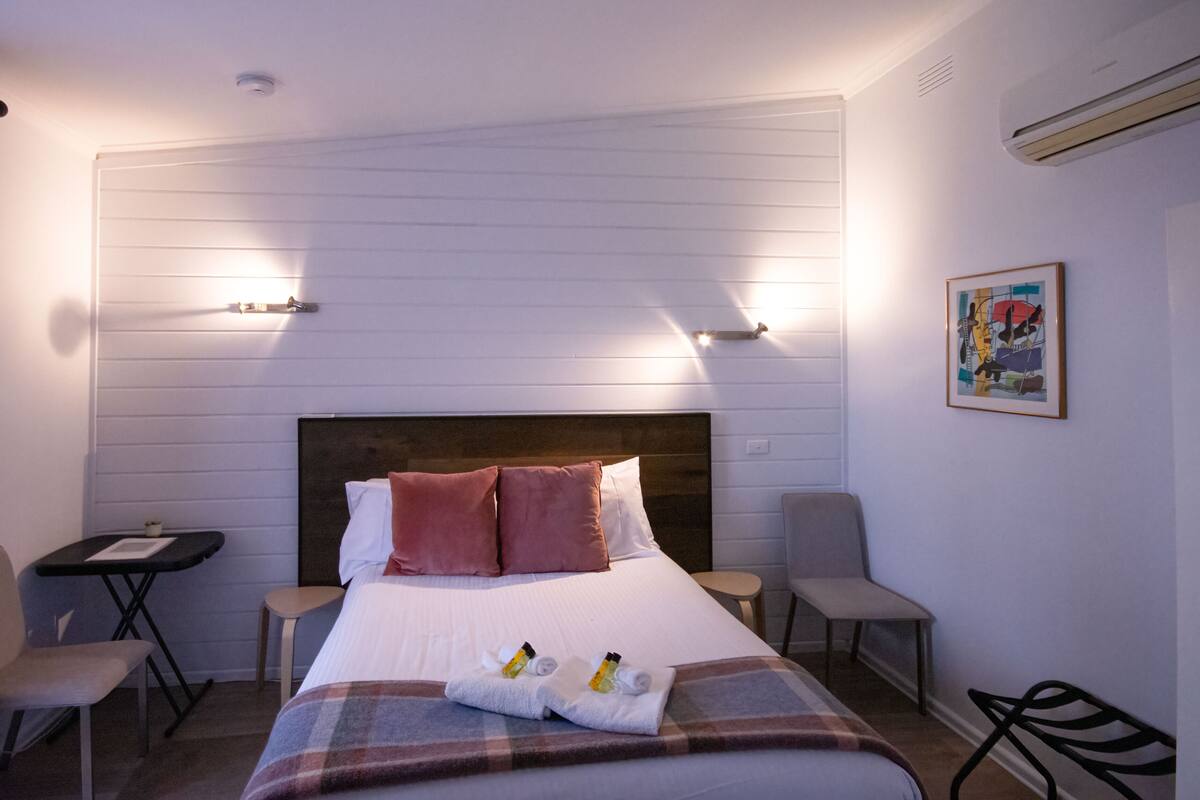 Una habitación de hotel con una cama, dos sillas, una mesita y un equipo de aire acondicionado.