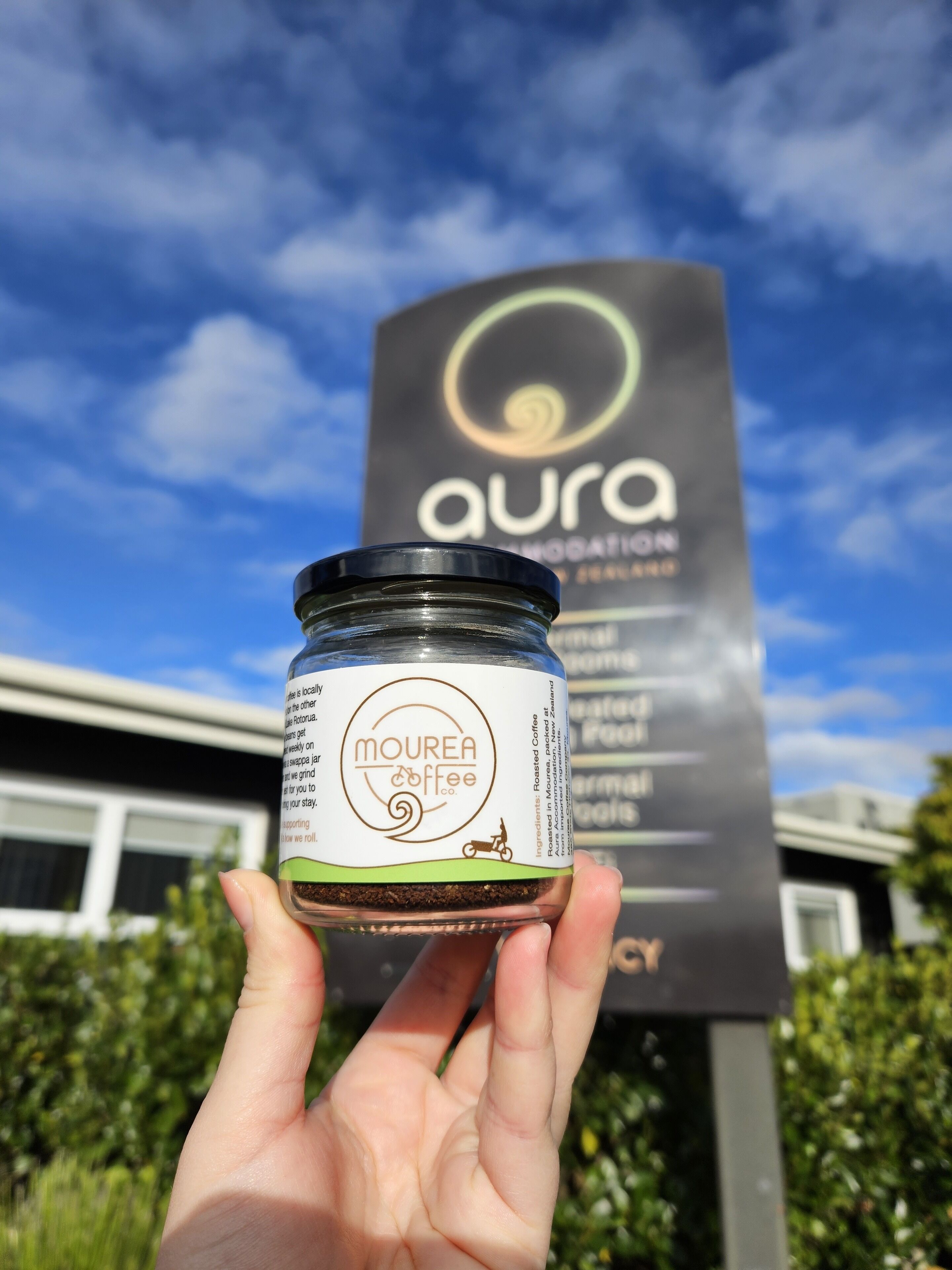 A hand holding a jar of Mourea Coffee in front of a sign for Aura.