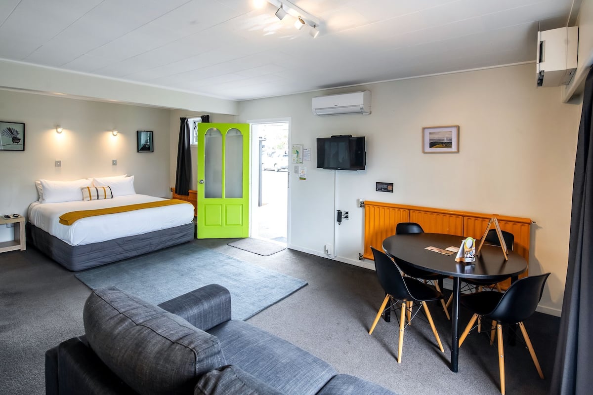 Una habitación de hotel con cama, un sofá gris, una mesa de comedor con sillas, una puerta verde y un televisor en la pared.