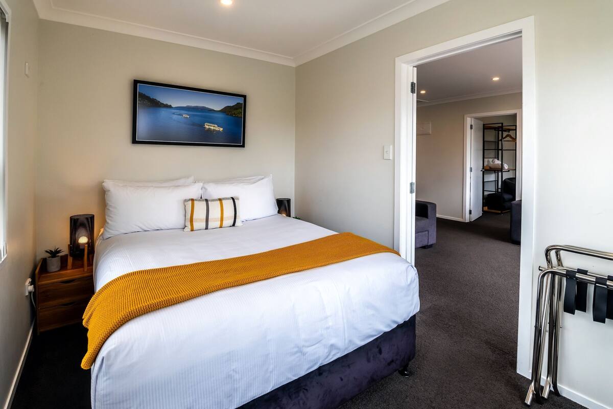 Habitación de hotel con una cama grande, una mesita de noche con lámpara, un cuadro en la pared y vistas al pasillo.