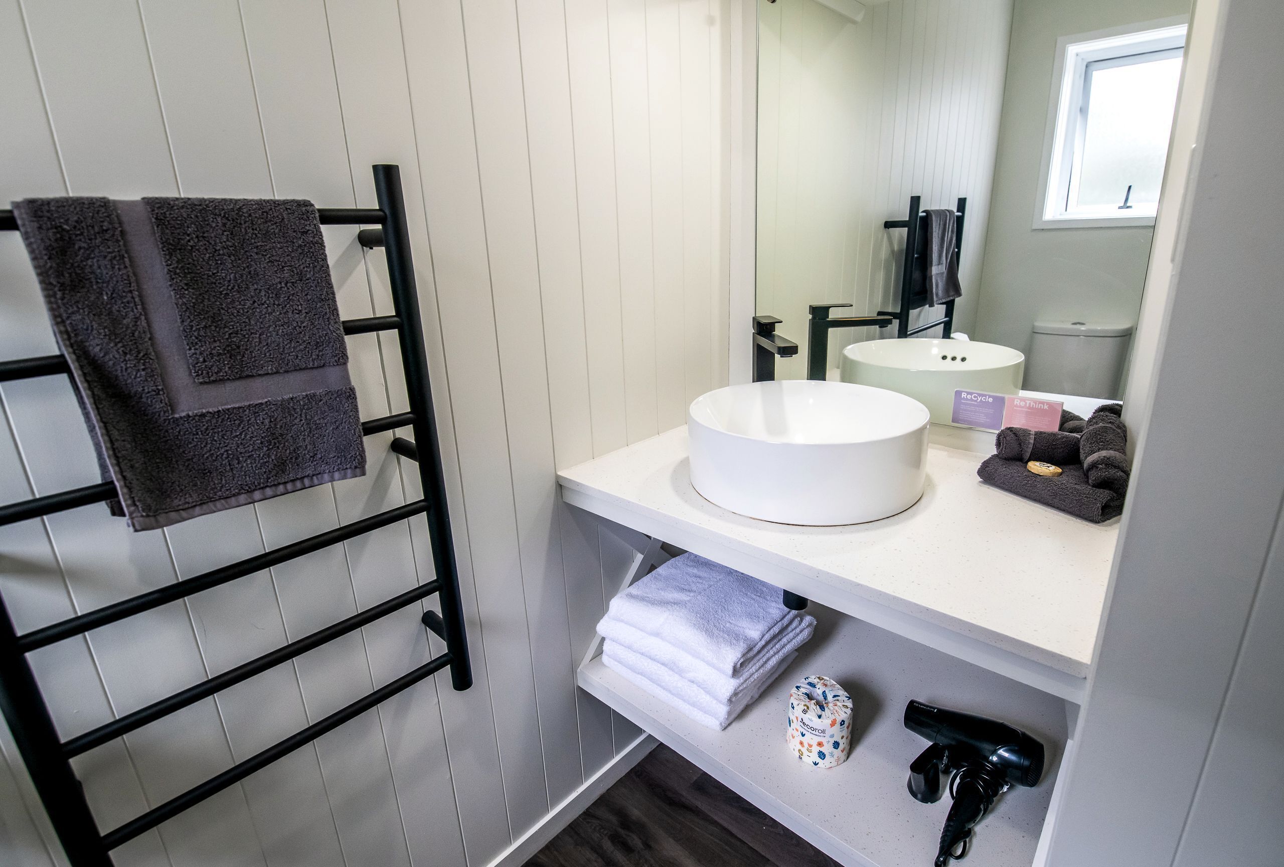 A bathroom with a white sink, a towel rack with folded towels, a mirror, and a window.