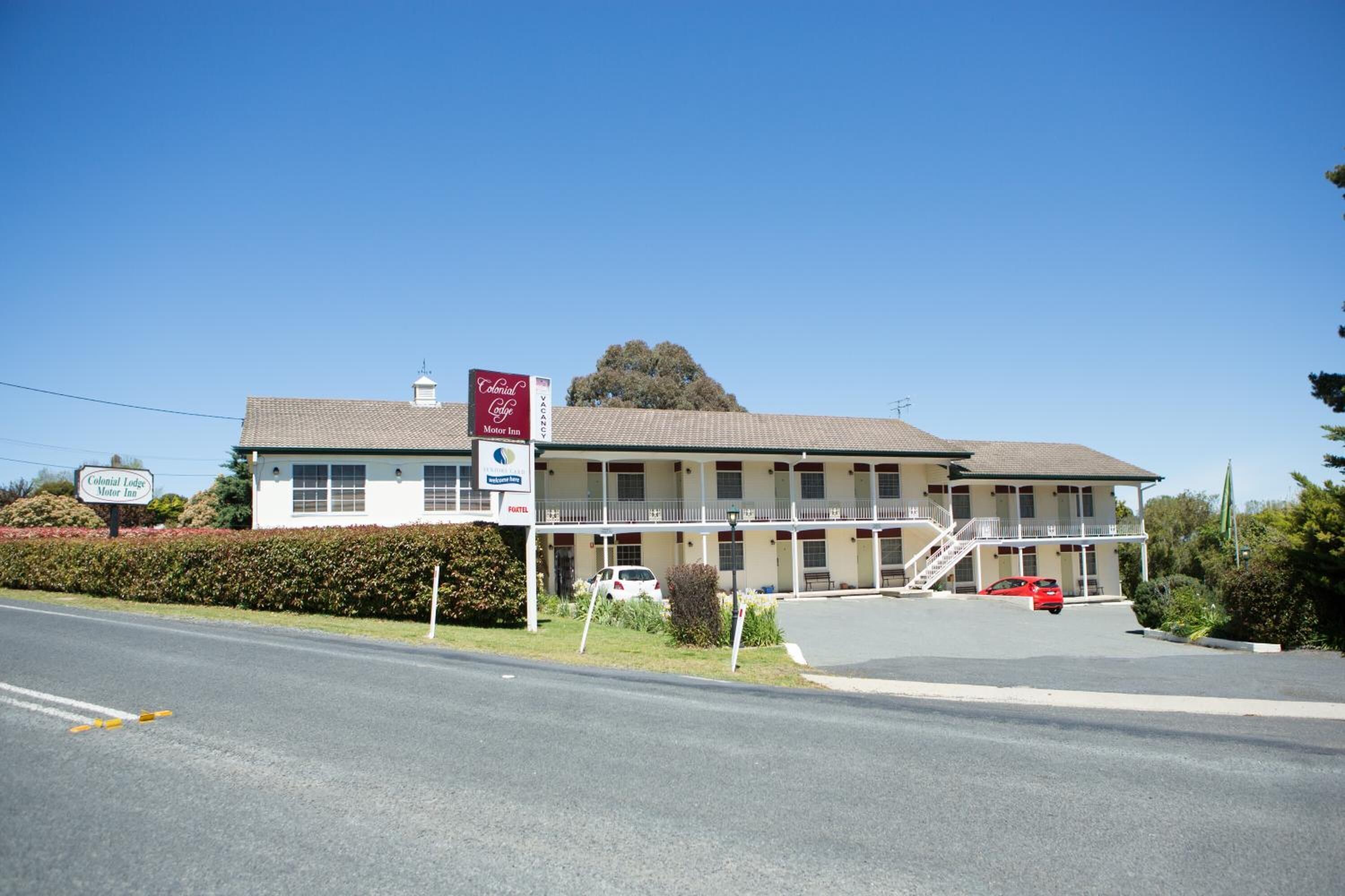 A Colonial Lodge Motor Inn with a two-story building, a covered front porch, and a parking lot with a red car.