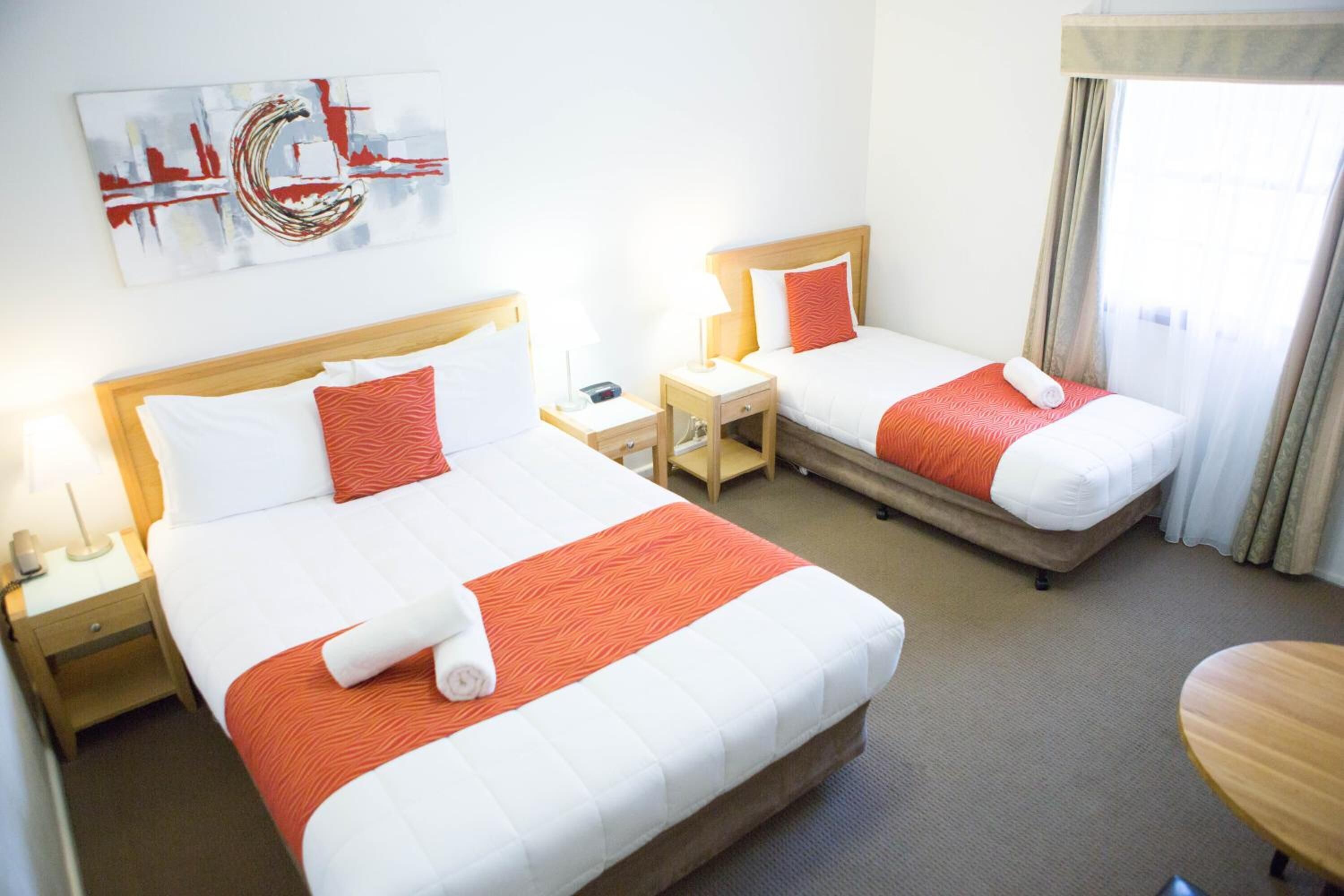 A hotel room with two beds, each with white and orange bedding, a nightstand with a lamp, and a painting on the wall.