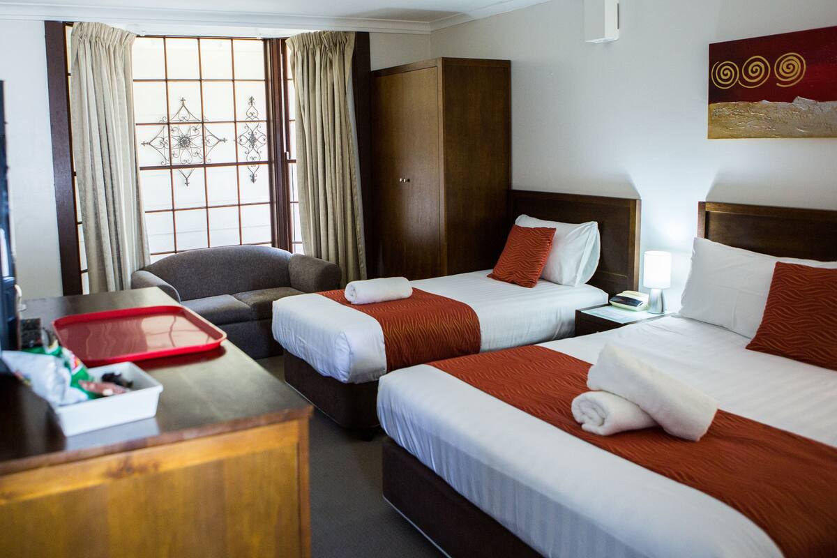 A hotel room with two beds, a wooden wardrobe, a desk with a red tray, and a window with curtains.