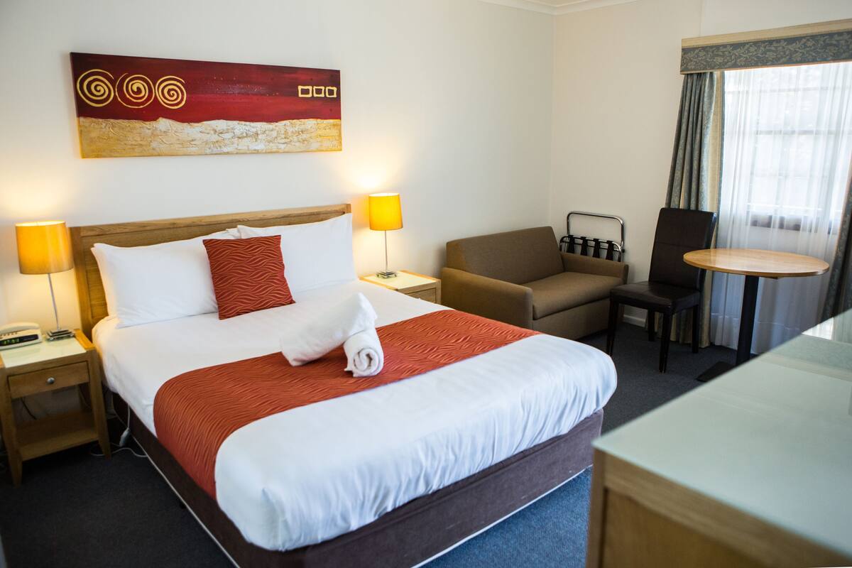 A hotel room with a large bed, two bedside tables, a chair, a small table, and a window with curtains.