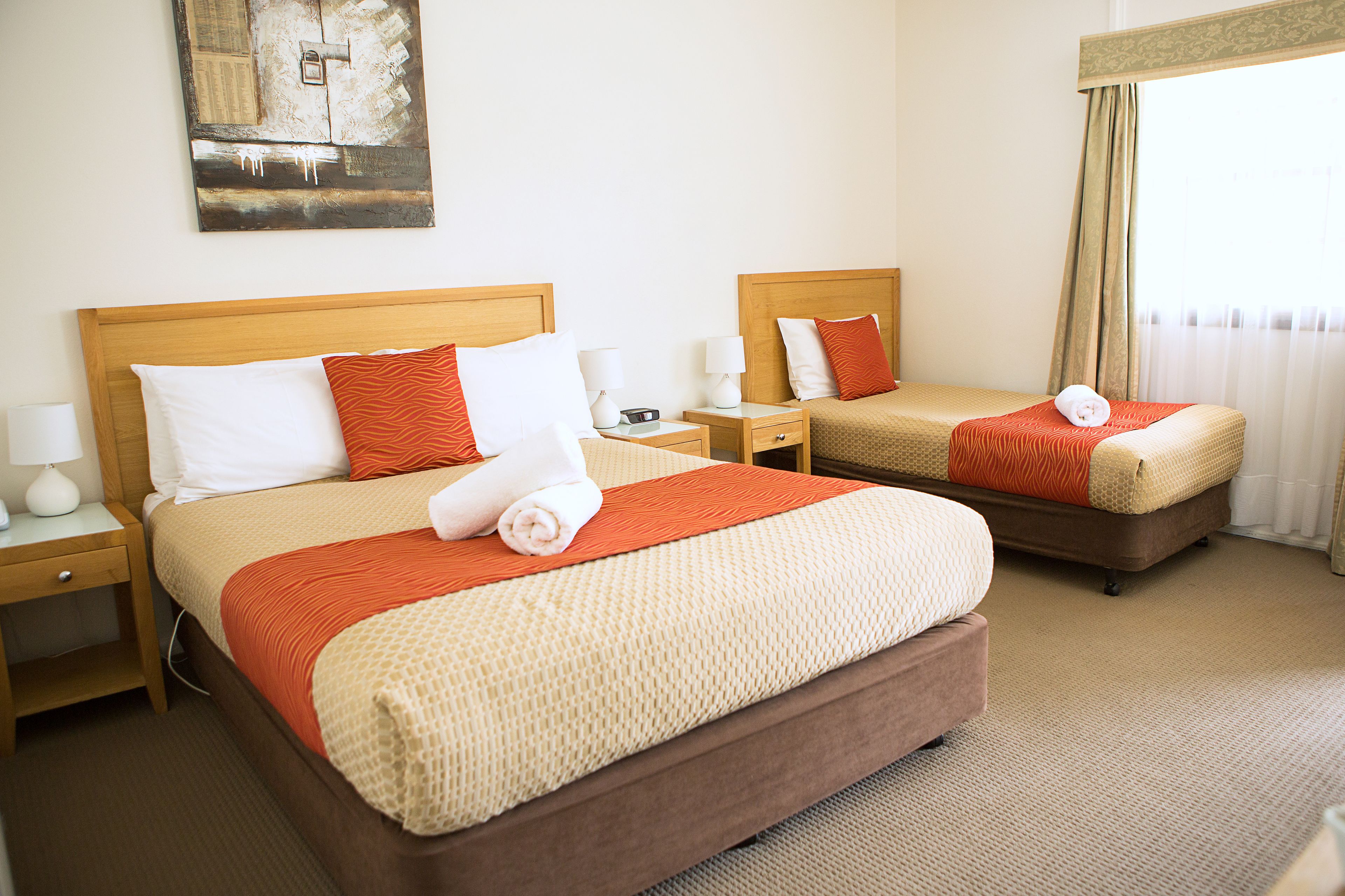 Quarto de hotel com duas camas, cada uma com cabeceira de madeira, almofadas brancas e laranja e um cobertor bege. Há mesas de cabeceira com abajures e um quadro na parede.