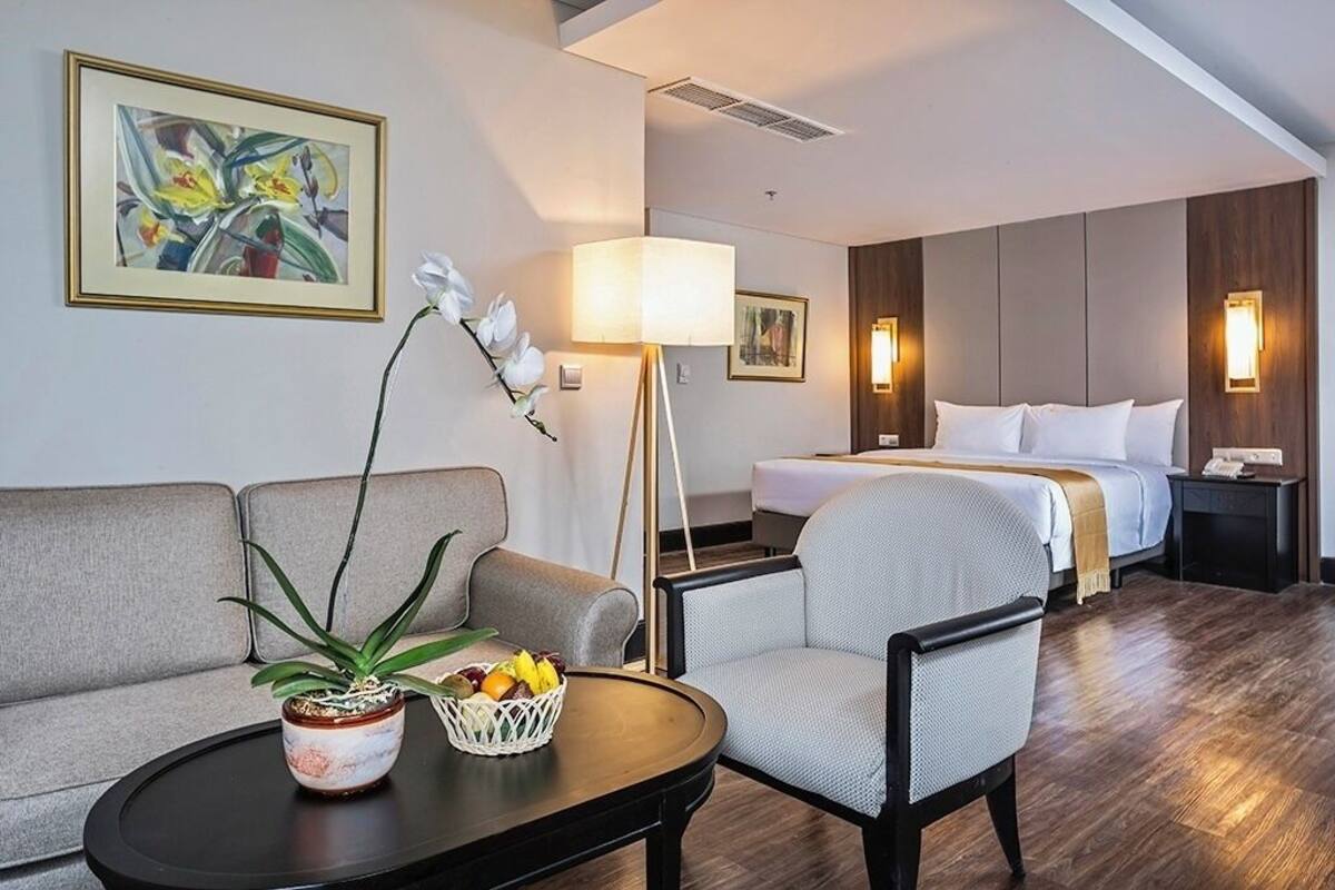 Une chambre d’hôtel avec un canapé, une table à manger agrémentée d’une plante et d’un panier de fruits, une lampe, un lit avec des oreillers et une table de chevet.