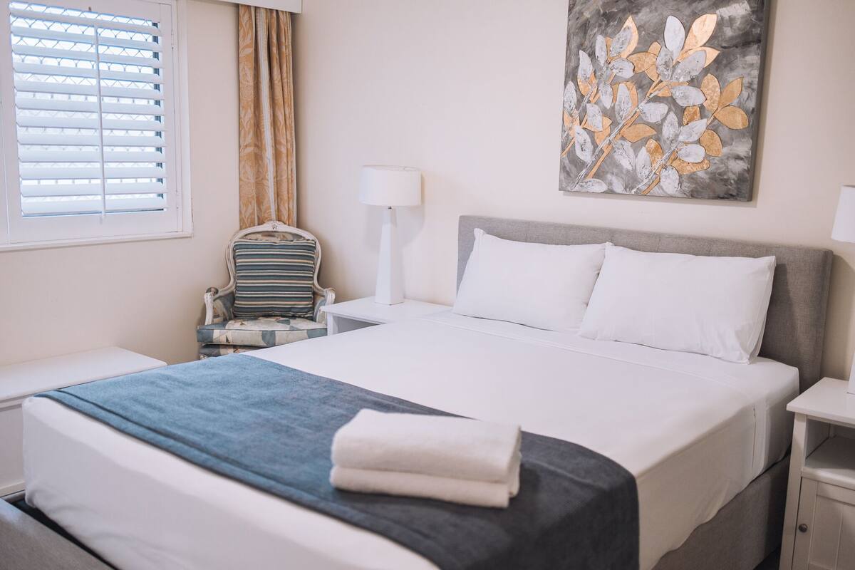 Un lit bien fait, avec une literie blanche et grise, un plaid bleu et une serviette blanche pliée. On aperçoit une table de chevet avec une lampe et un téléphone. Une chaise avec un coussin à rayures est placée à côté du lit. Une fenêtre avec des stores et un rideau est visible.