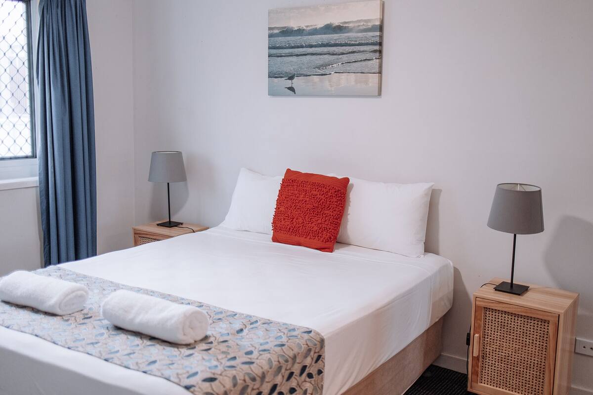 Un lit bien fait, avec des oreillers blancs et rouges, une couvre-lit bleue et blanche à motifs, et deux lampes de chevet posées sur des tables de nuit en bois.