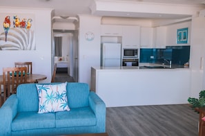 A modern living room with a blue sofa, a wooden dining table, and a kitchen area with white cabinets and a microwave. - Surfers Horizons Resort (Palm Beach)