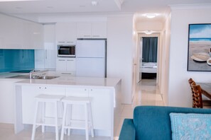 A modern kitchen with white cabinets, a built-in oven, and a large island with bar stools. There is a dining area with a blue sofa and a wooden chair. - Surfers Horizons Resort (Palm Beach)