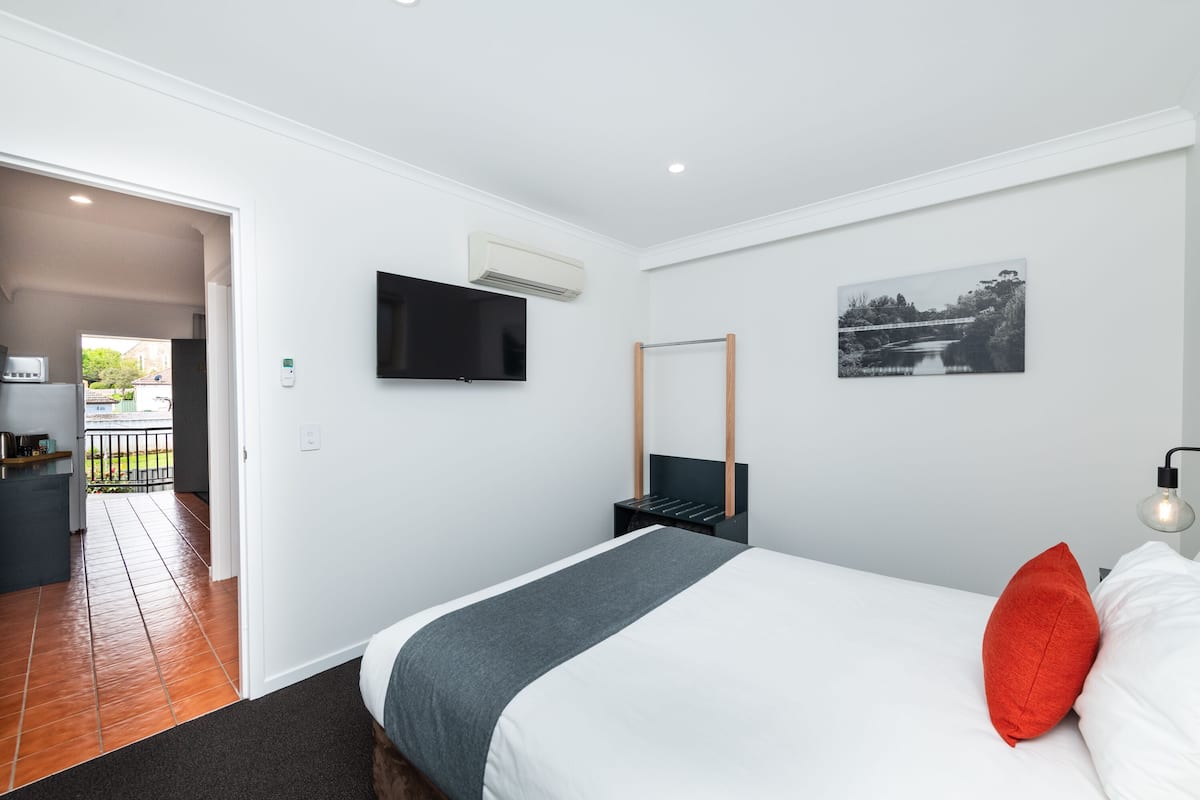 Una habitación de hotel moderna con una cama, un televisor montado en la pared, un cuadro y una mesita de noche con una lámpara.
