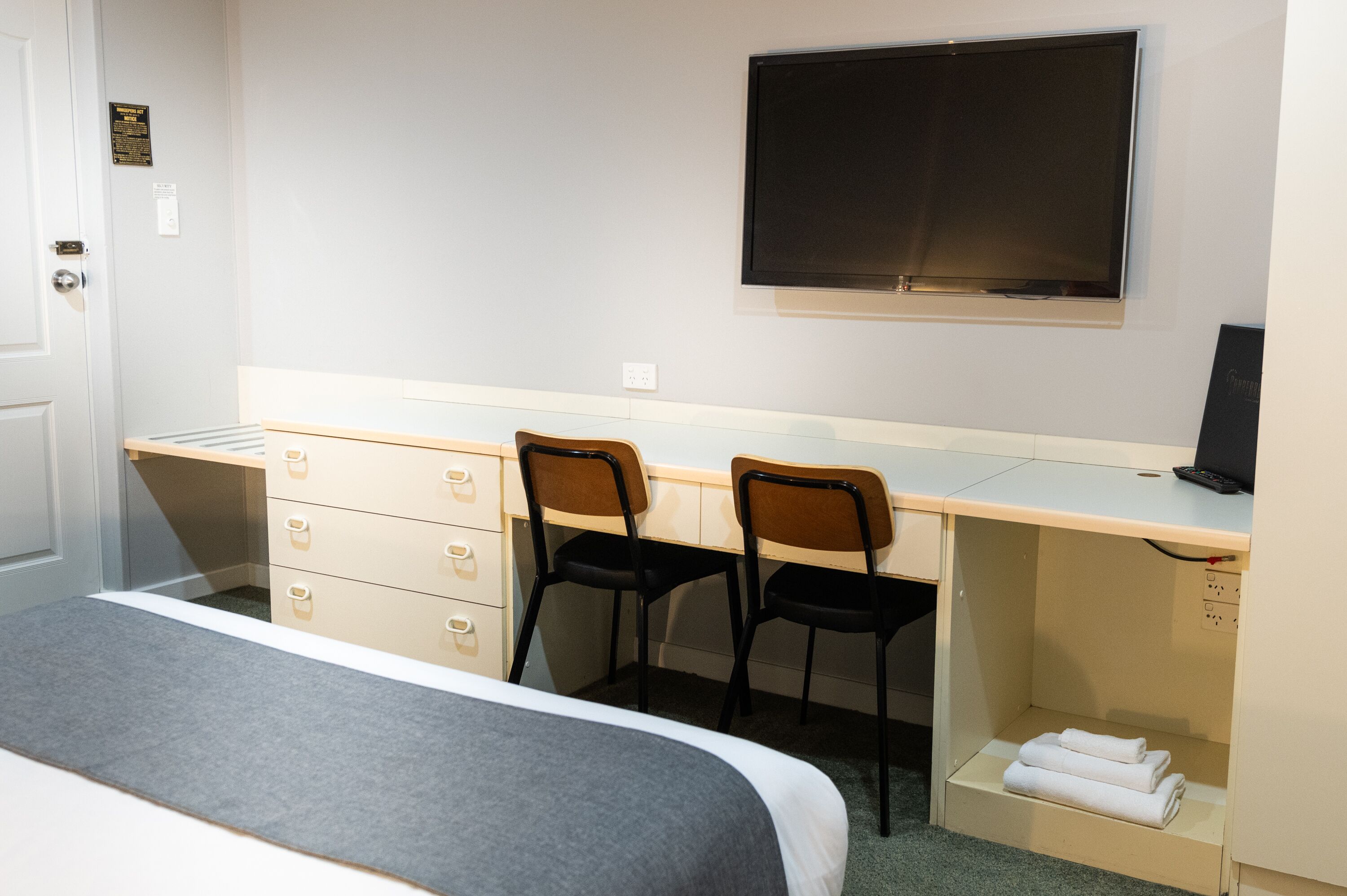 A hotel room with a bed, a desk with two chairs, a flat-screen TV, and a small shelf with towels.