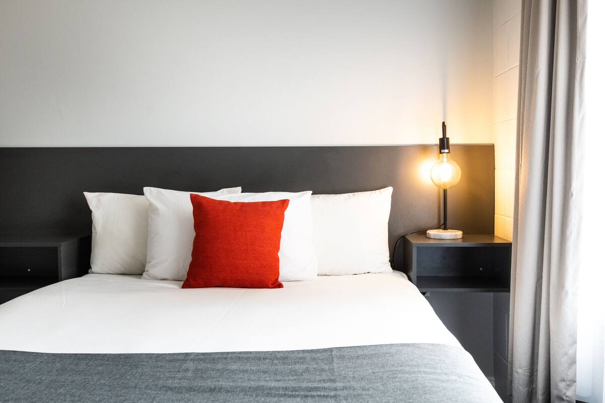 Una cama bien hecha con almohadas blancas y rojas, una mesita de noche con lámpara y una ventana con cortinas.