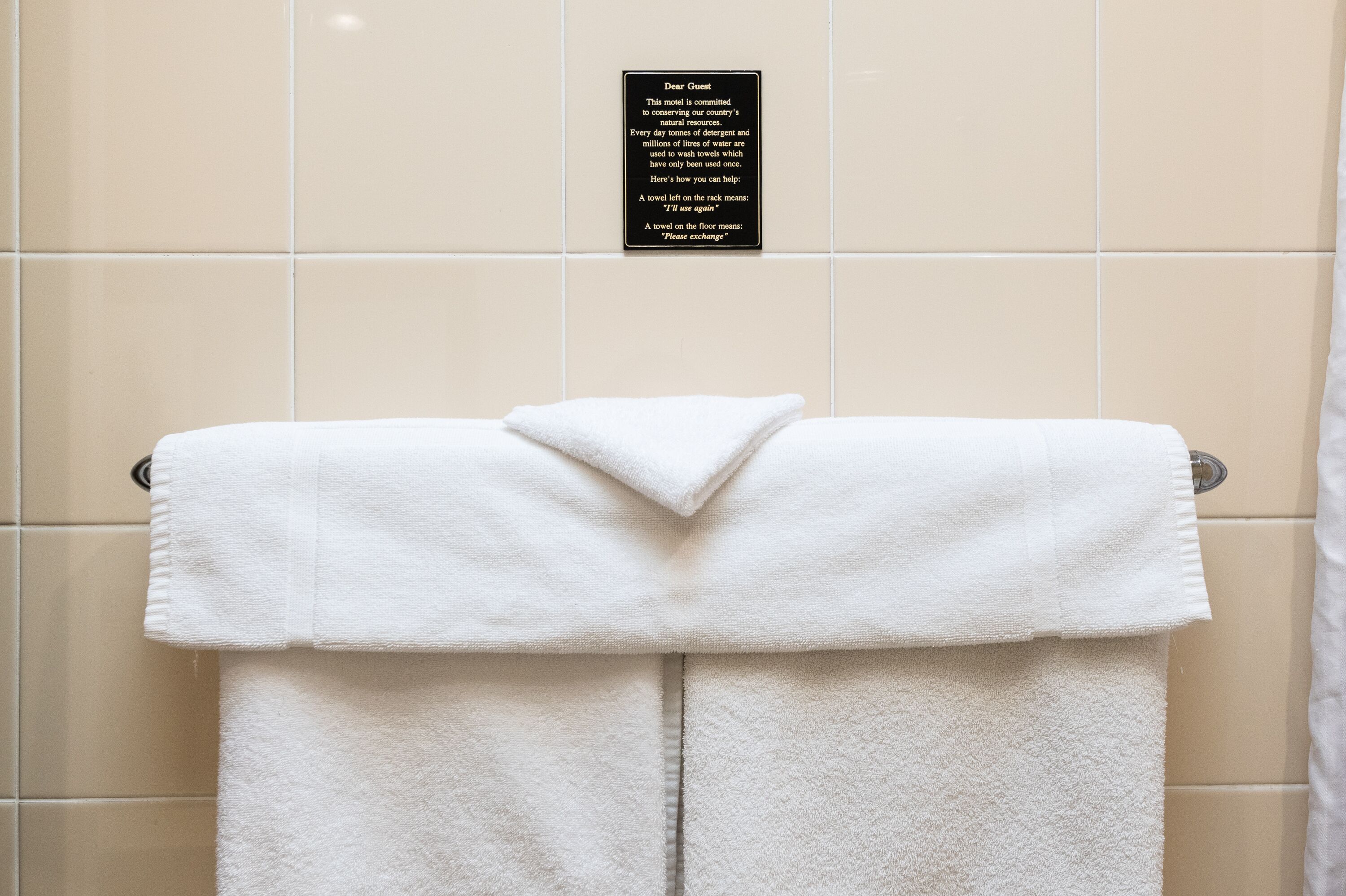 A white towel hanging on a metal towel rack in a bathroom with beige tiles. A sign above the towel explains the hotel's commitment to conserving water and provides instructions for towel reuse.