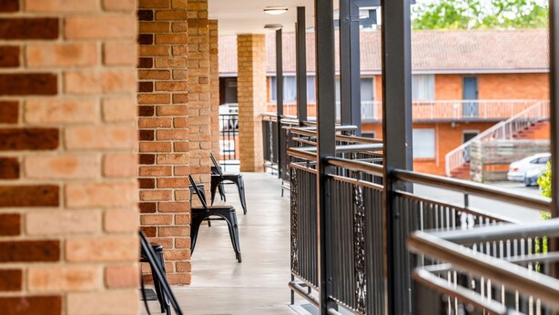 A corridor with brick walls, black chairs, and a railing overlooking a street with buildings and cars. - River Motel (Queanbeyan East)