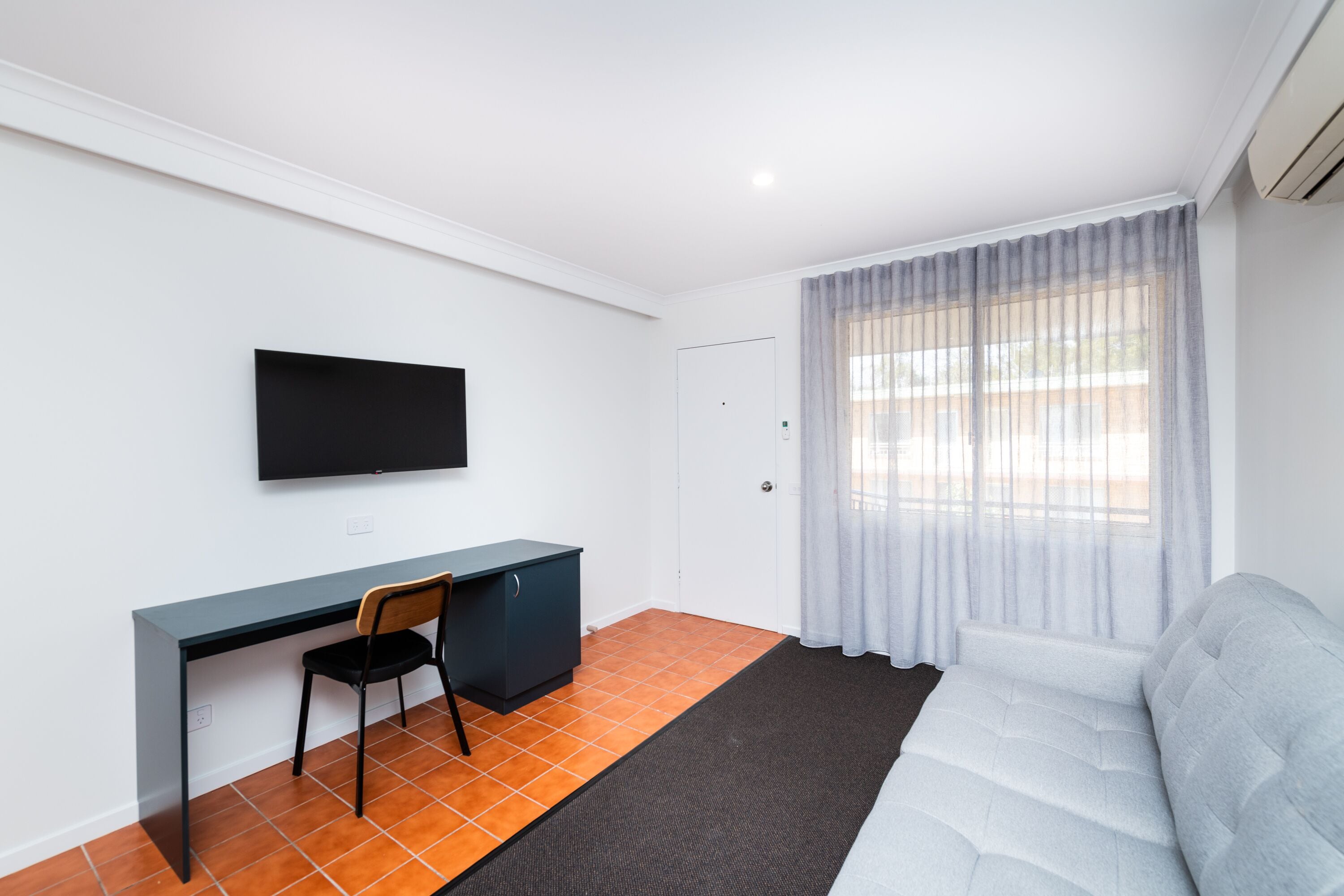 Una habitación con cama, escritorio con silla, televisor de pantalla plana en la pared y ventana con cortinas traslúcidas.
