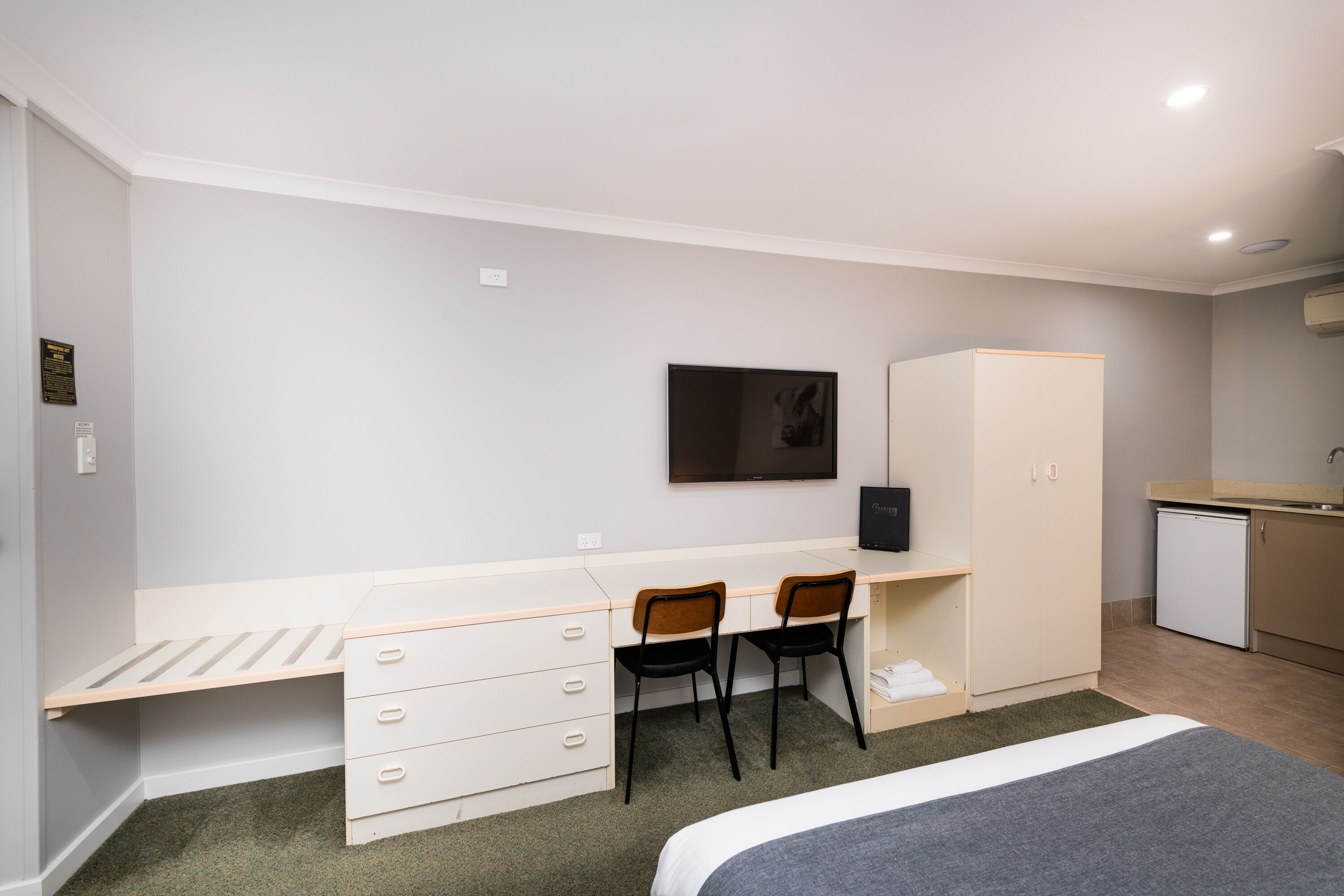 Un dormitorio con cama, escritorio, dos sillas, armario, televisor y una pequeña kitchenette.