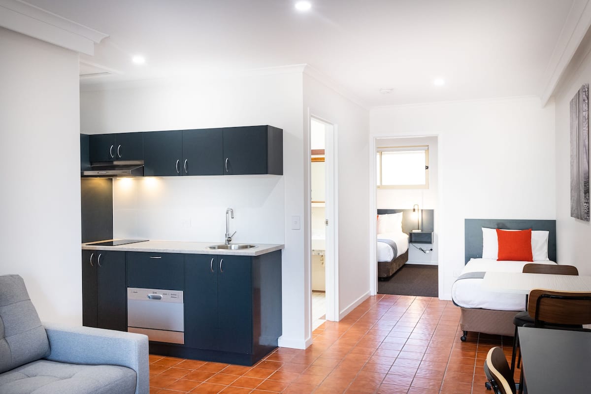 Una habitación de hotel compacta con kitchenette, una cama con almohada roja y un pequeño comedor.