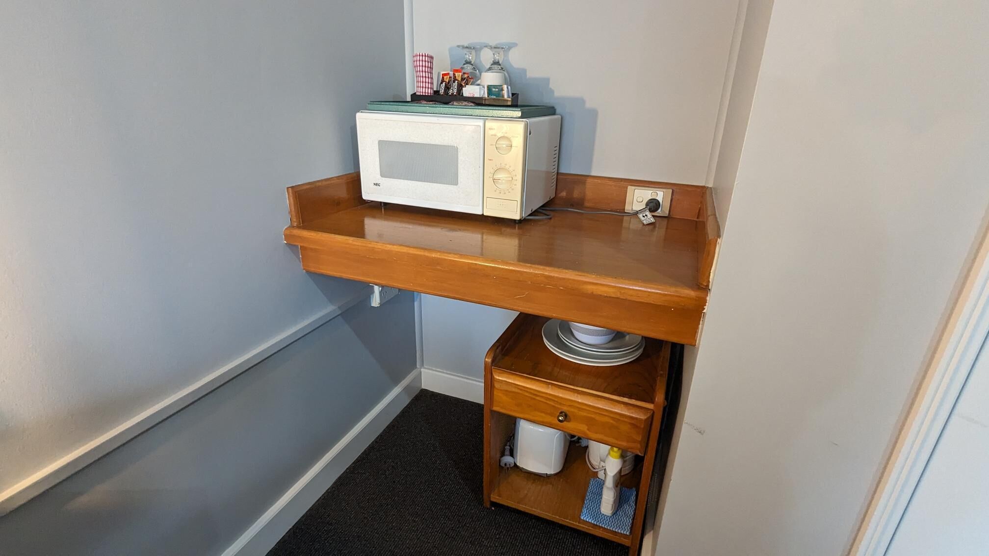 A compact kitchenette with a microwave, a wooden shelf, and a small drawer.