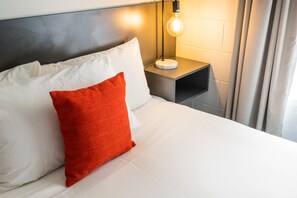Una cama bien hecha con almohadas blancas y rojas, una mesita de noche con una lámpara y una cortina en la ventana.