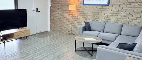 Una sala moderna con un sofá gris, una mesita redonda pequeña, un televisor de pantalla plana sobre un mueble de madera y una pared de ladrillo con un cuadro enmarcado.