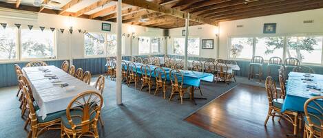A large indoor dining area with multiple tables and chairs, wooden beams on the ceiling, and large windows allowing natural light.