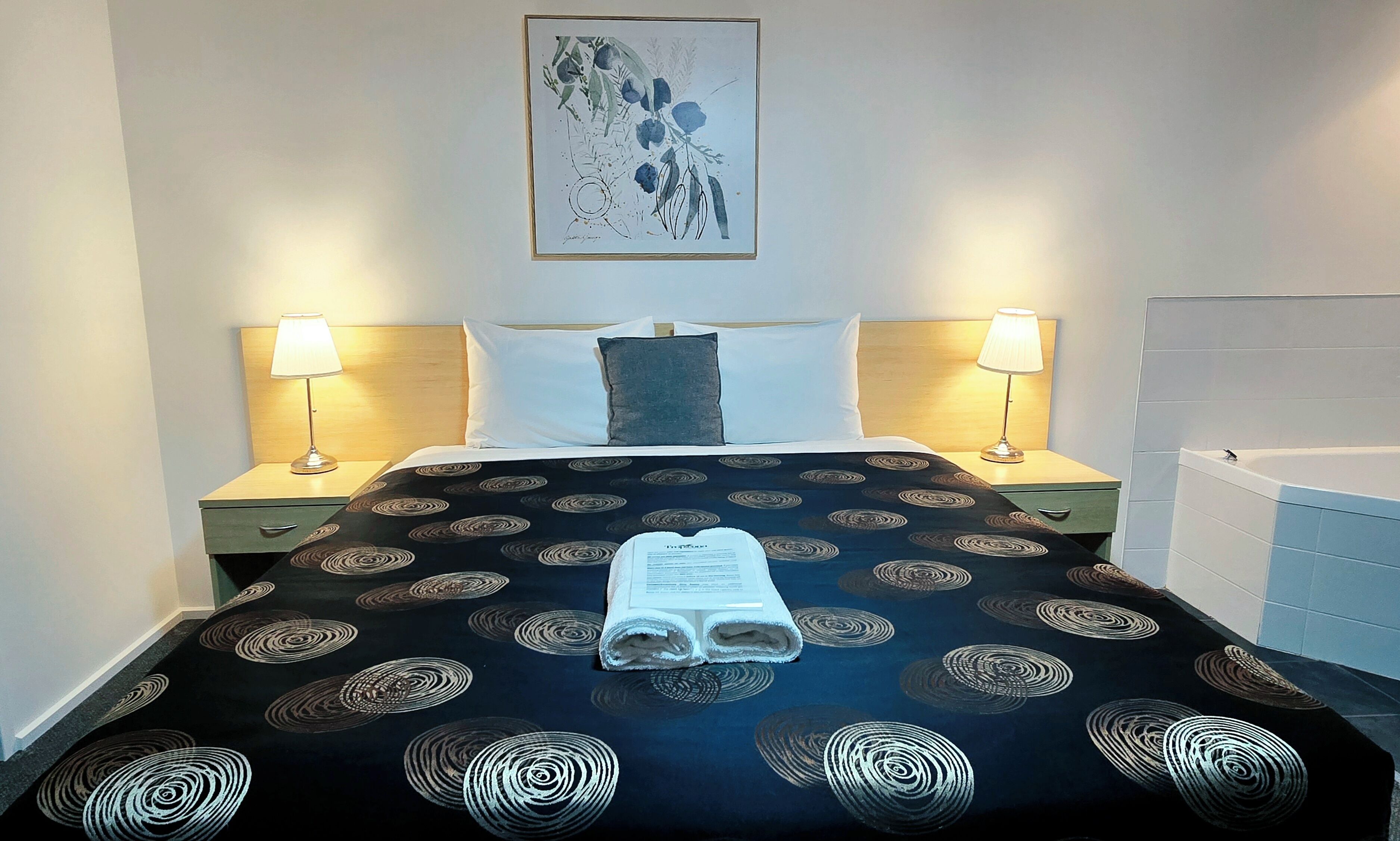 A hotel room with a bed, two bedside lamps, a headboard, a framed artwork, and a bathroom with a bathtub.