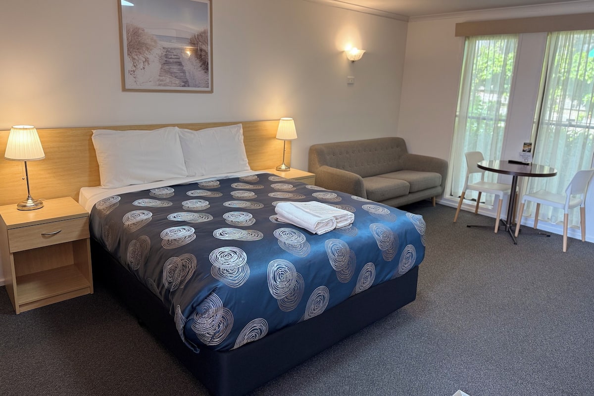 Habitación de hotel con cama, dos lámparas de noche, un sofá, una mesa con sillas y una ventana con cortinas.