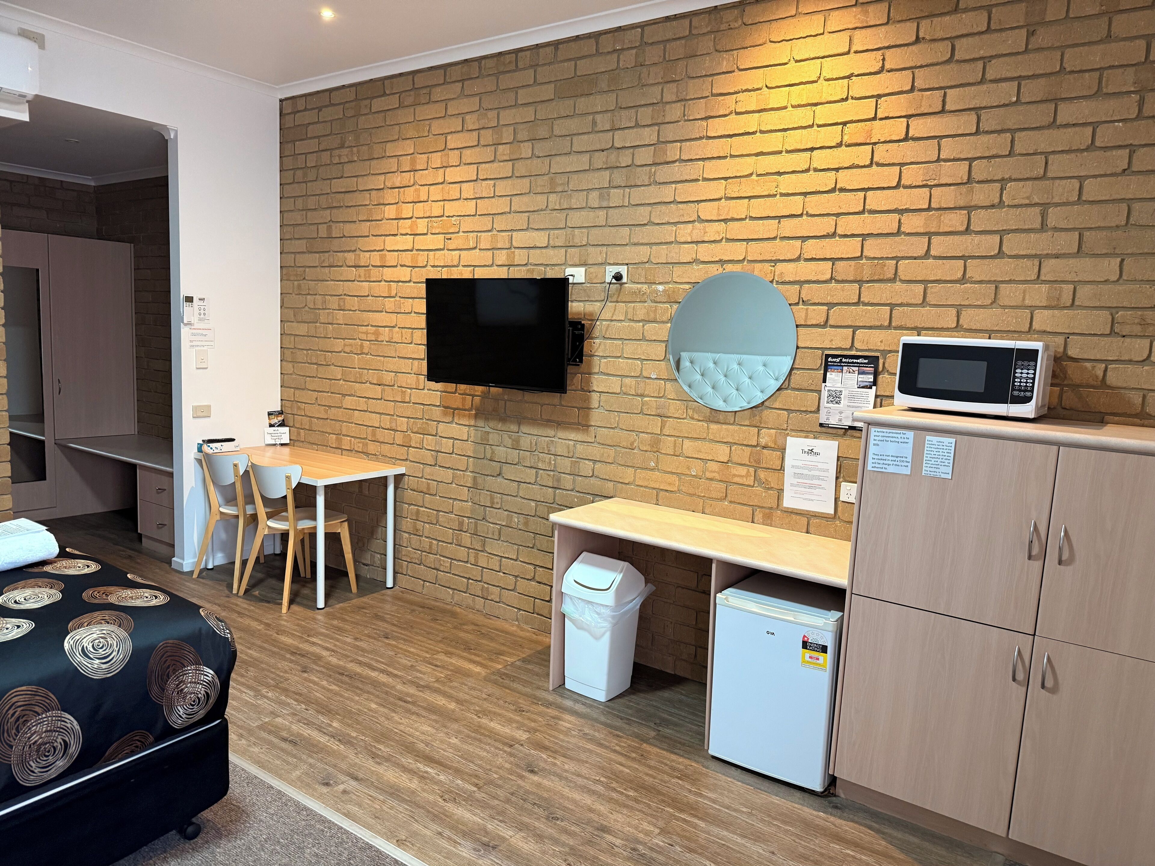 Habitación compacta con pared de ladrillo, televisión de pantalla plana, microondas, refrigerador pequeño, escritorio con sillas y sofá.