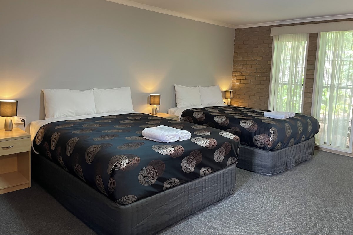 Habitación de hotel con dos camas, cada una con una colcha estampada, lámparas de noche y un ventanal con cortinas translúcidas.