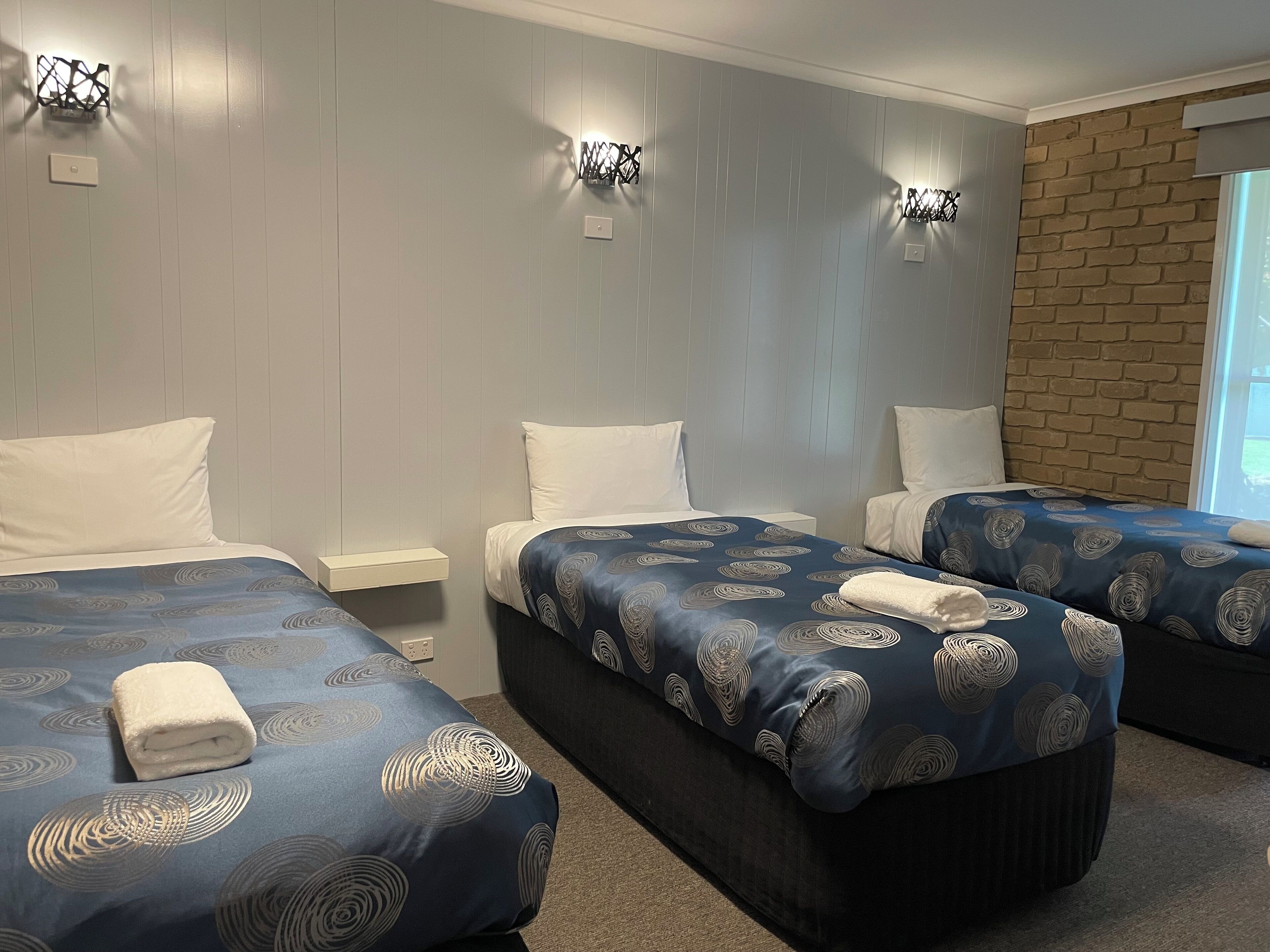 Habitación de hotel con dos camas, cada una con una colcha azul estampada y almohadas blancas. Hay lámparas de pared y una pared de ladrillo decorativa.