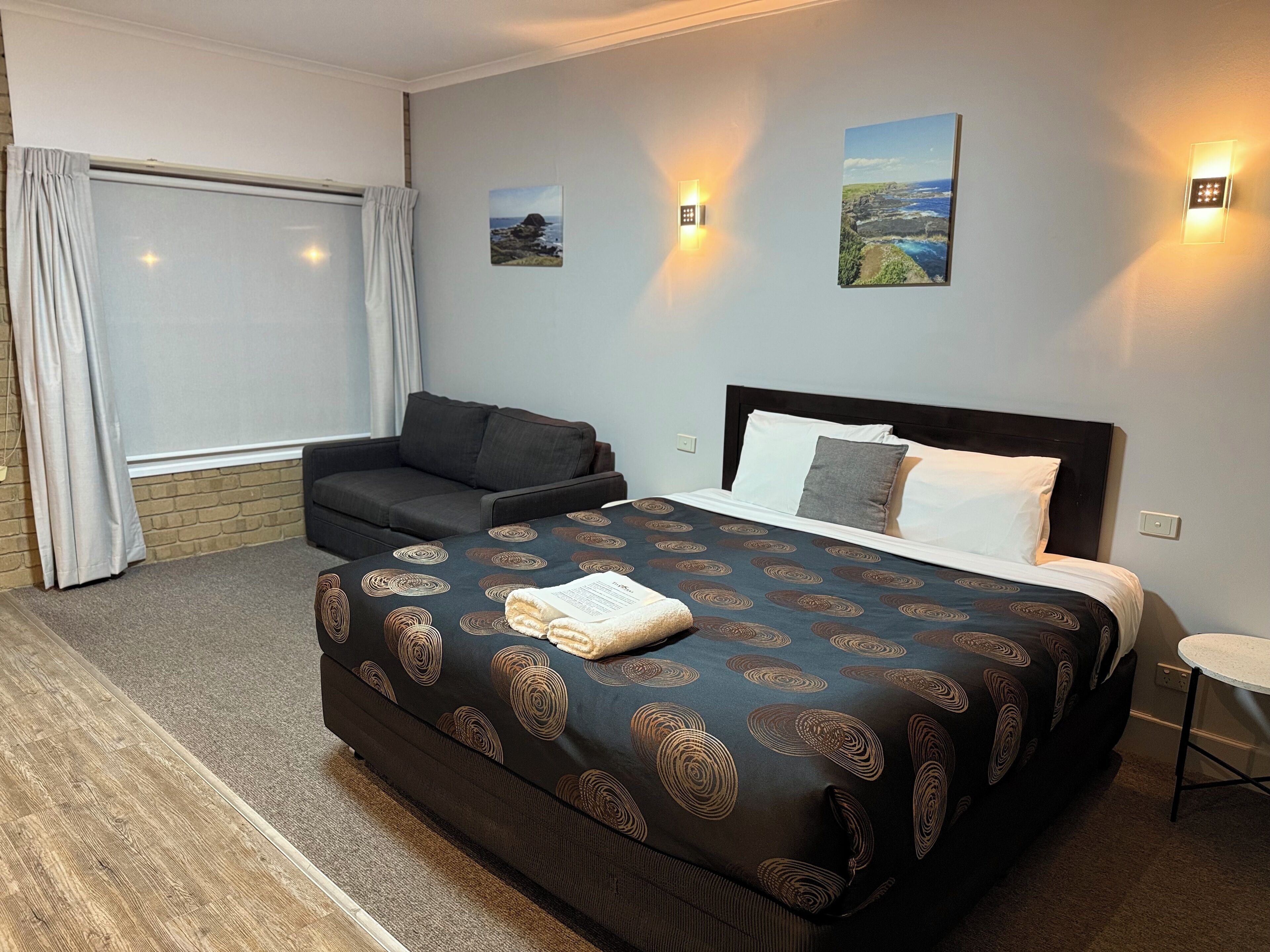 A hotel room with a large bed, a sofa, a small table, and two wall-mounted lights.