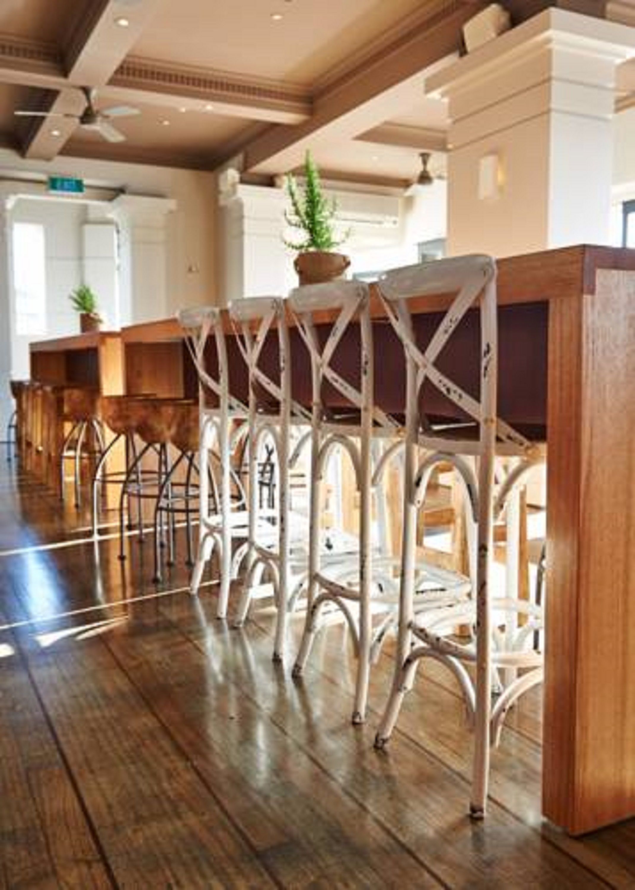 A row of white metal bar stools with a crisscross design, wooden tables, and a polished wooden floor in a well-lit room with a high ceiling and ceiling fans.