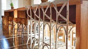 A row of white metal bar stools with a crisscross design, wooden tables, and a polished wooden floor in a well-lit room with a high ceiling and ceiling fans.
