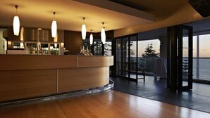 A modern interior with a wooden reception desk, pendant lighting, and a view of the ocean through large glass doors. - Cottesloe Beach Hotel (Cottesloe)