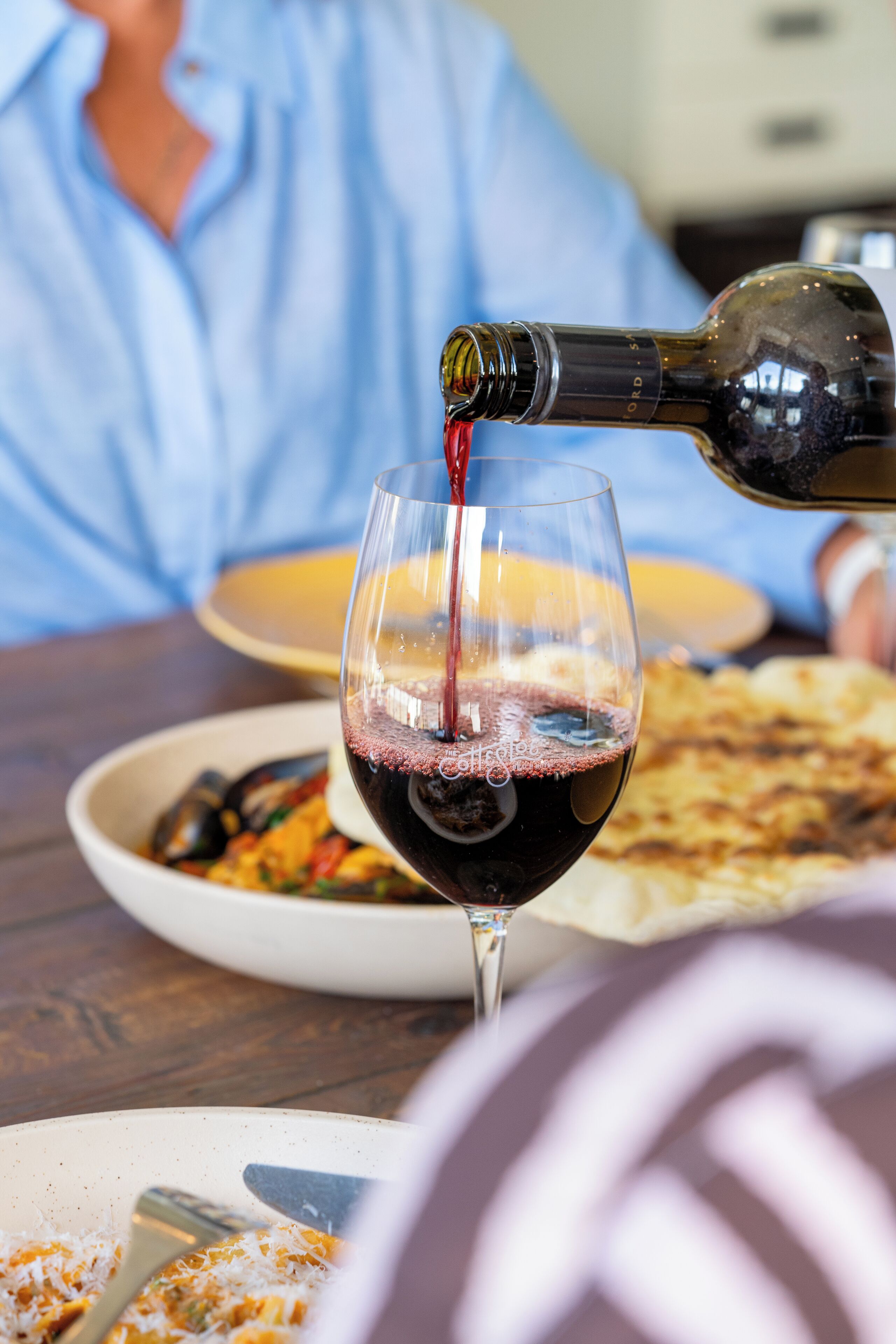 A dining table with a wine bottle pouring into a glass, a plate of food, and a person in the background.