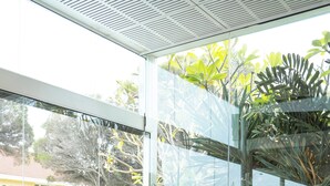 A modern living room with a wooden floor, white sofas with cushions, a coffee table, and a glass wall with a view of greenery and buildings. - Cottesloe Beach Hotel (Cottesloe)