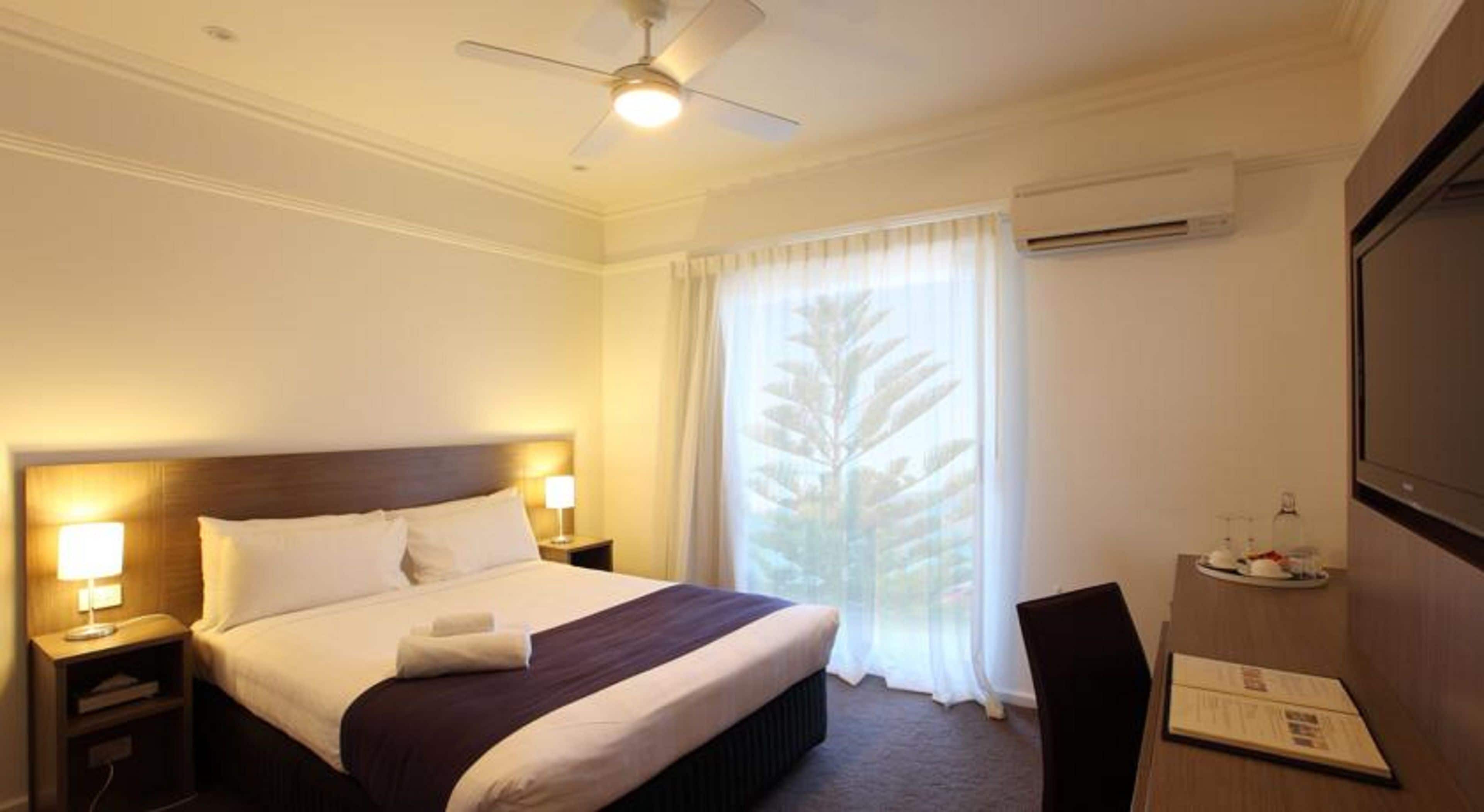 A hotel room with a bed, a desk, a chair, a ceiling fan, and a window with curtains.
